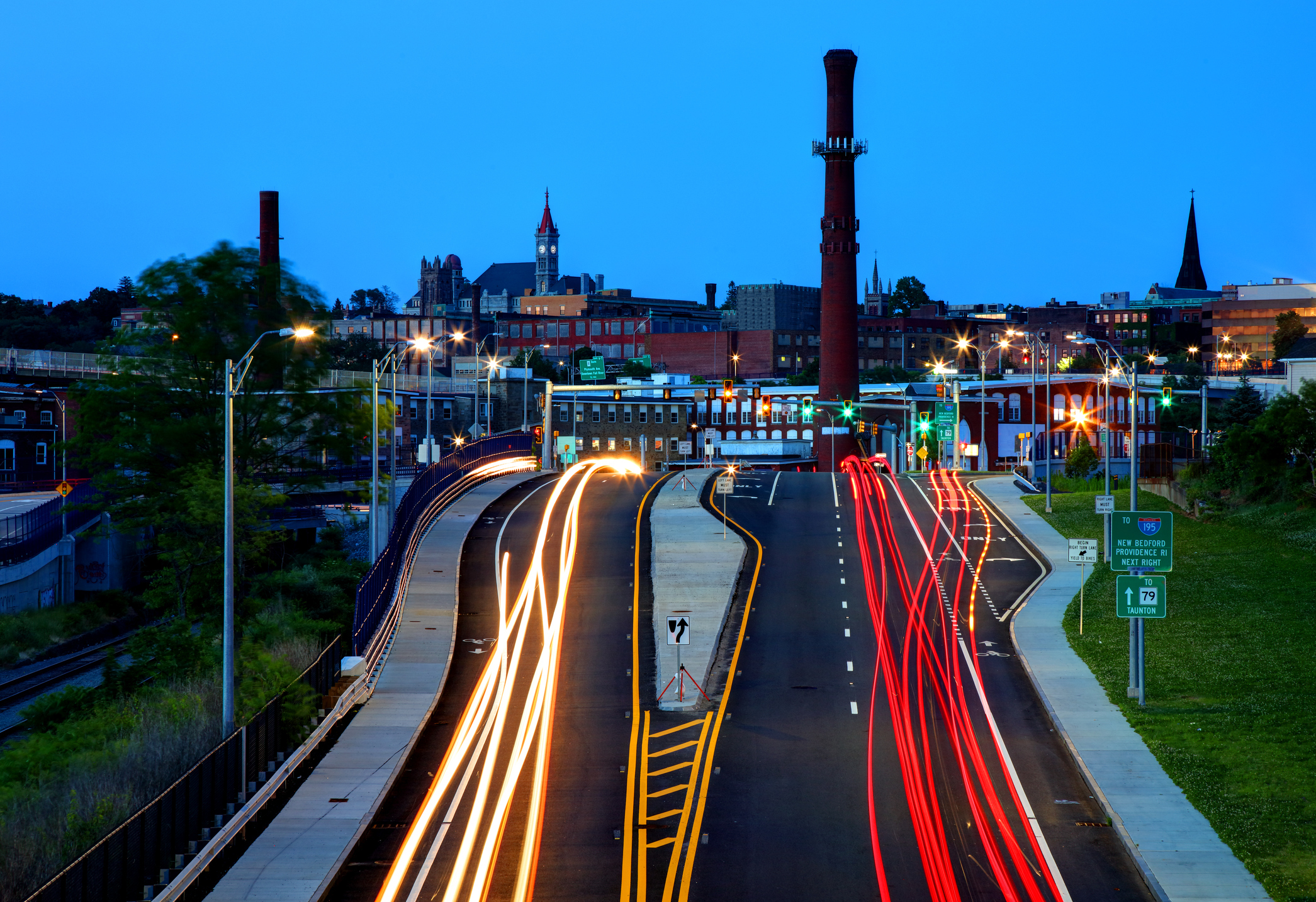 Downtown Fall River, Massachusetts in the evening, showing the town's smokestack, church steeples, and streaks of traffic on the highway, with the city skyline in the back.