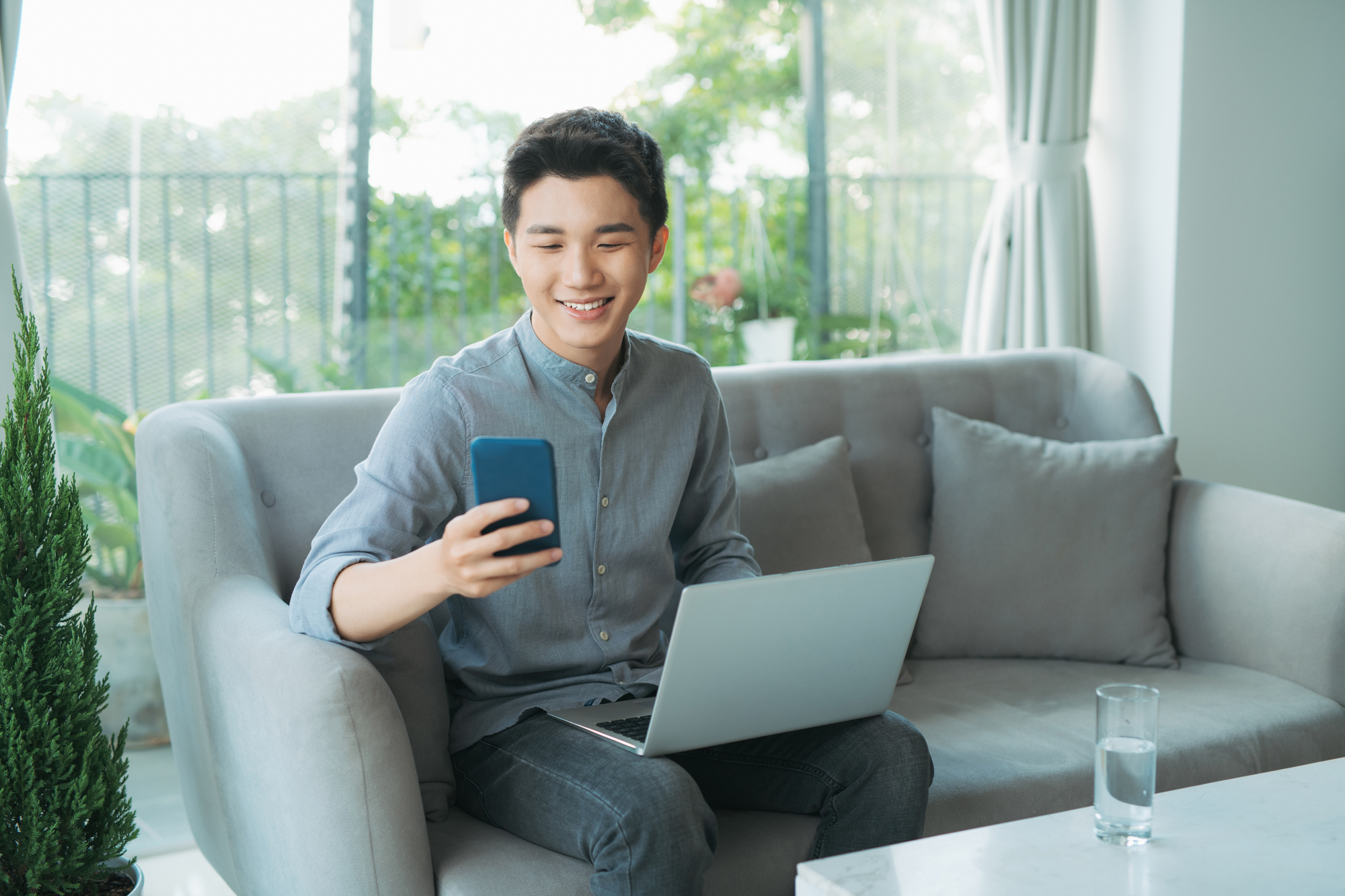 Young man smiling while using smartphone and laptop on couch