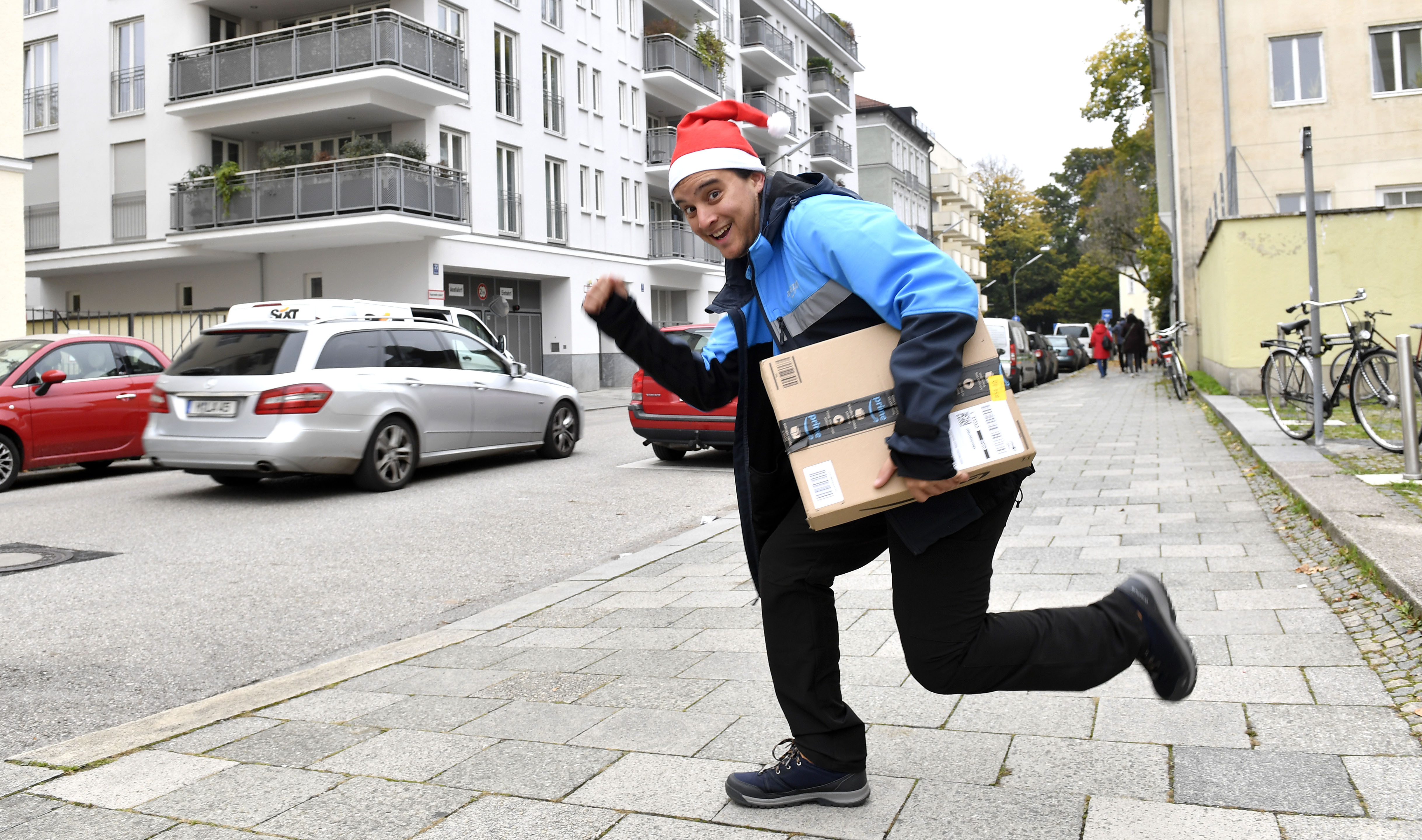 Ein junger Mann in Amazon-Zusteller-Kleidung mit roter Weihnachtszipfelmütze: Er hält ein Amazon Paket in der Hand.