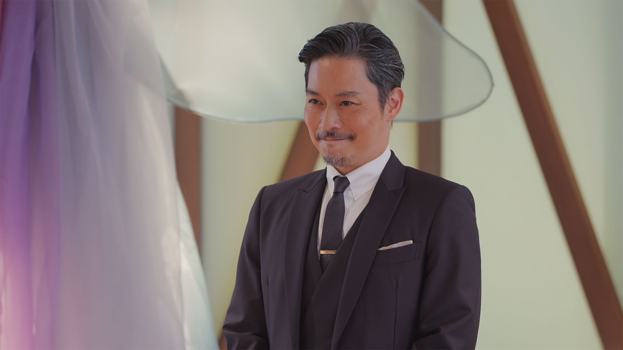 室内で三つ揃いスーツの男性が立ち、微笑んでいる。背後は白い壁に木の柱が見え、紫色の薄い布がひるがえっている。