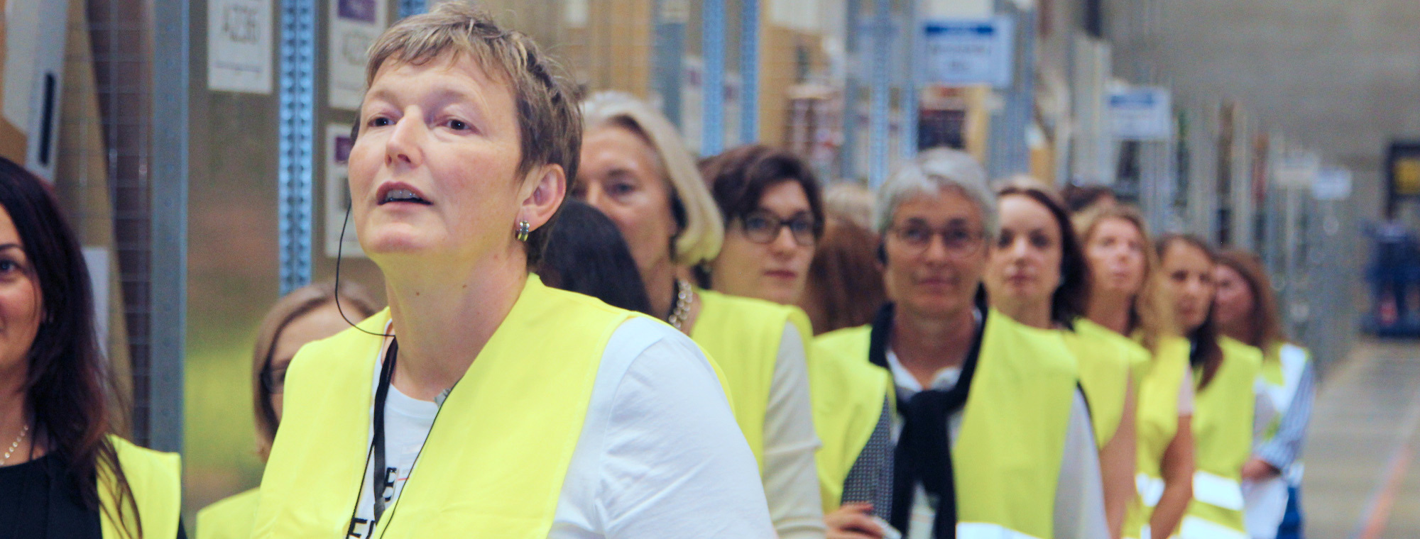 Mehrere Frauen mittleren Alters in gelben Sicherheitswesten stehen hintereinander. Im Hintergrund sind man die Regale des Logistikzentrums.