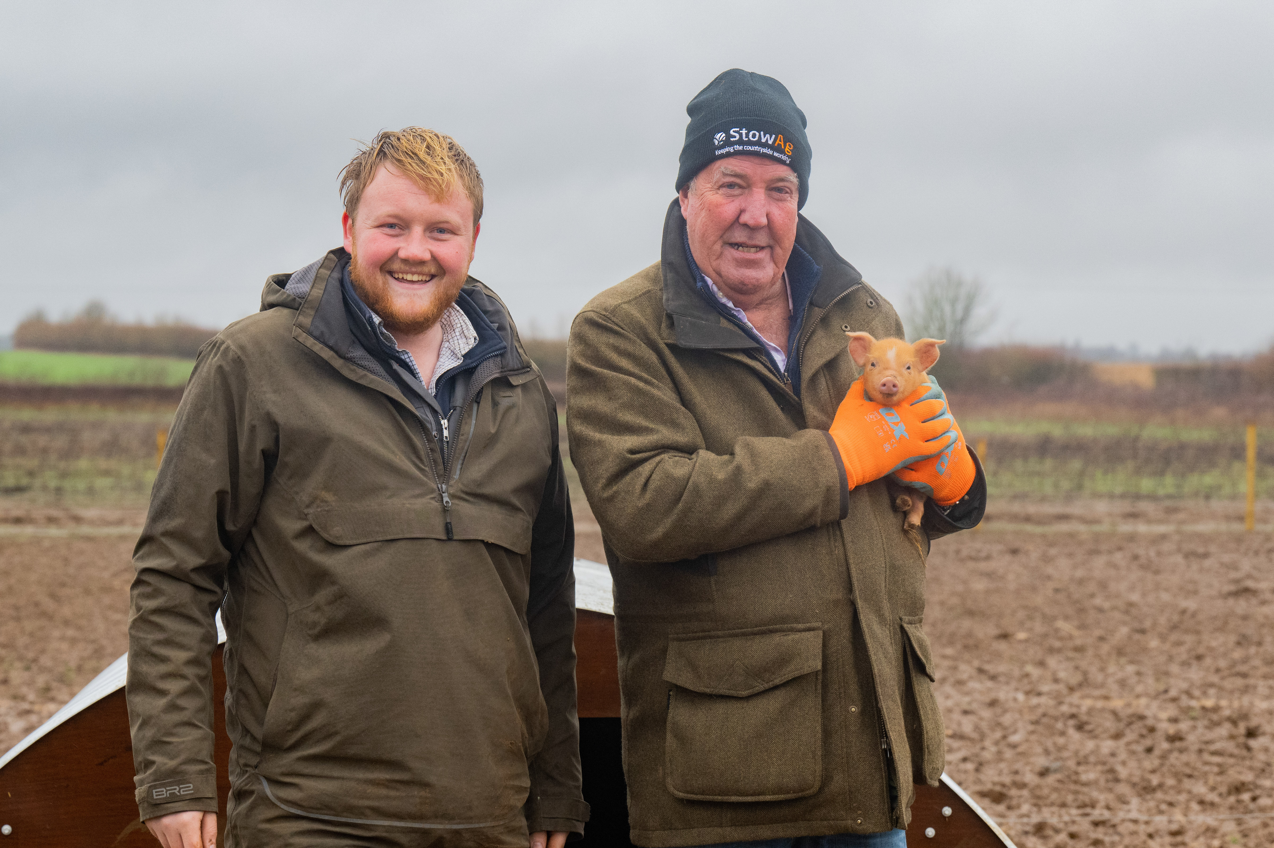 Two men holding a small pig with a farm in the foreground