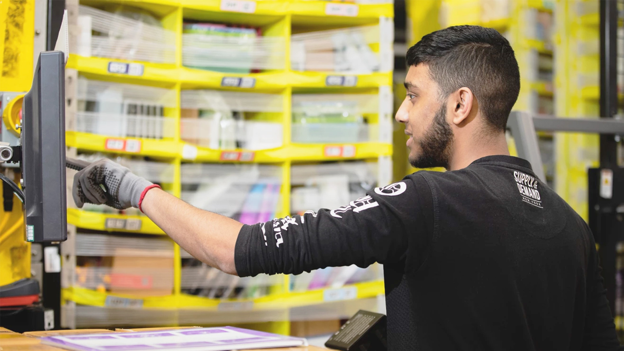 A man wearing a black shirt and work gloves taps a screen while working in an Amazon fulfillment center.