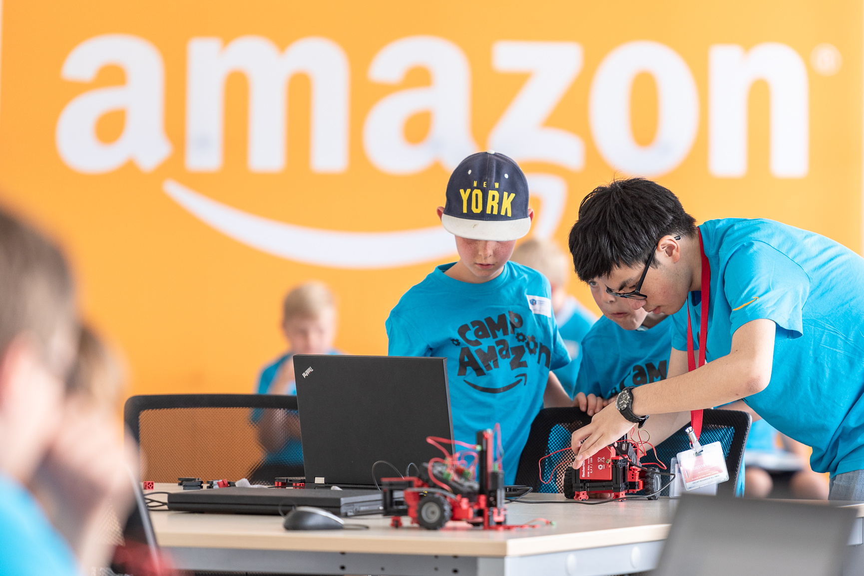 2 Kinder in blauen T-Shirts mit der Aufschrift "Camp Amazon" basteln an einem Tisch an kleinen Gabelstaplern