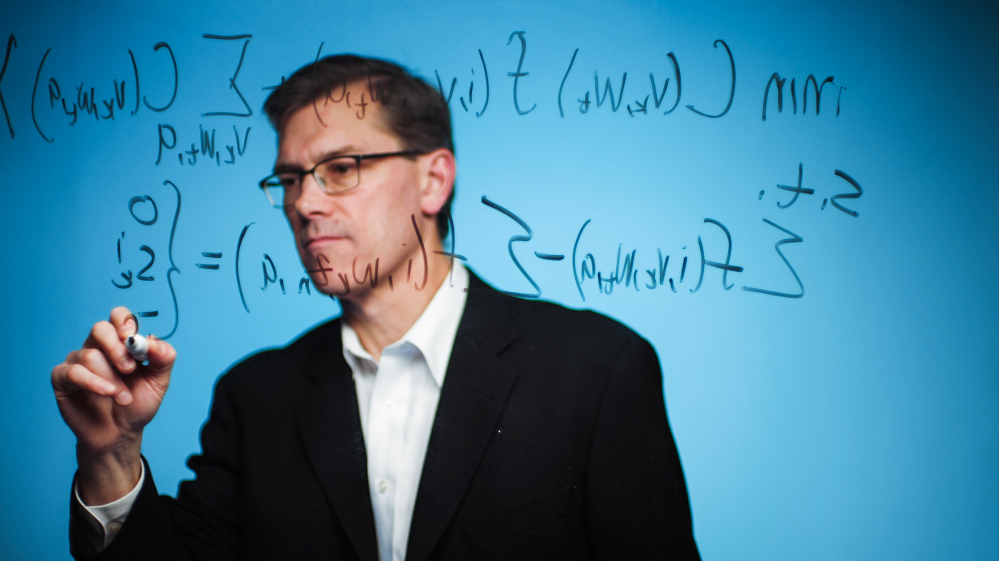 A man wearing a blazer and a button-down shirt writes a complicated equation.