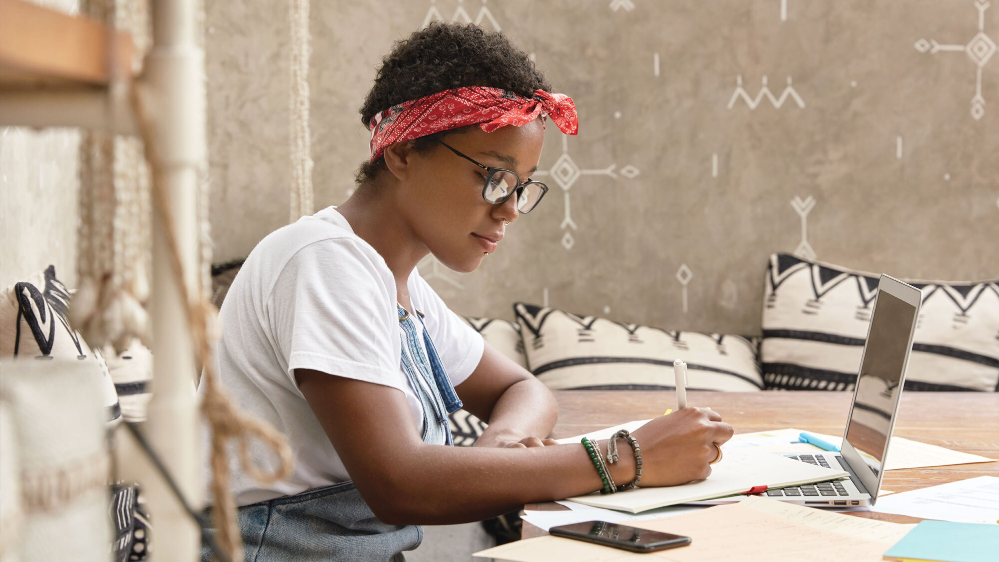 A photo of a person sitting at a desk, writing on a pad of paper that is placed on top of their laptop device.