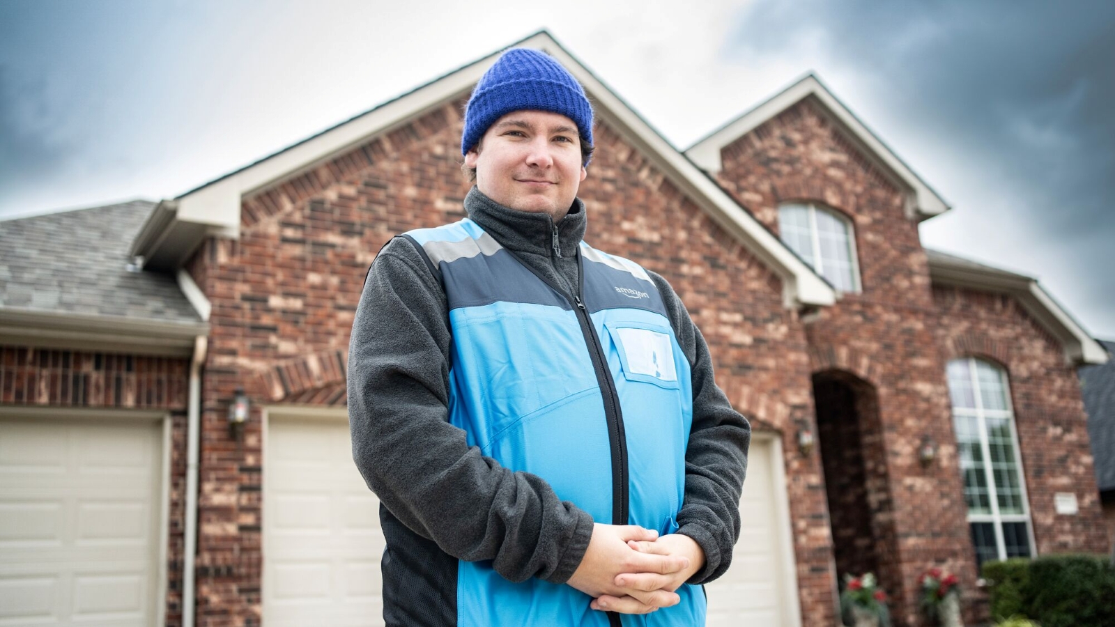Amazon delivery associate Kyle Sullivan, wearing a blue vest and beanie, smiles in front of brick home.
