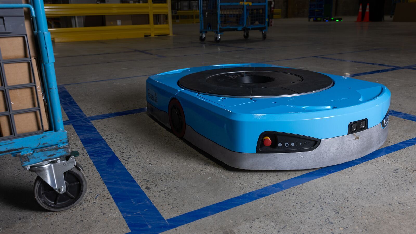 An image of the Titan robot. It is in an Amazon fulfillment center on a concrete surface, sitting next to a rolling rack with boxes inside. The robot is a plate-like bot with a blue shell and black features, including a round plate on top, a camera on the front, and several sensors.