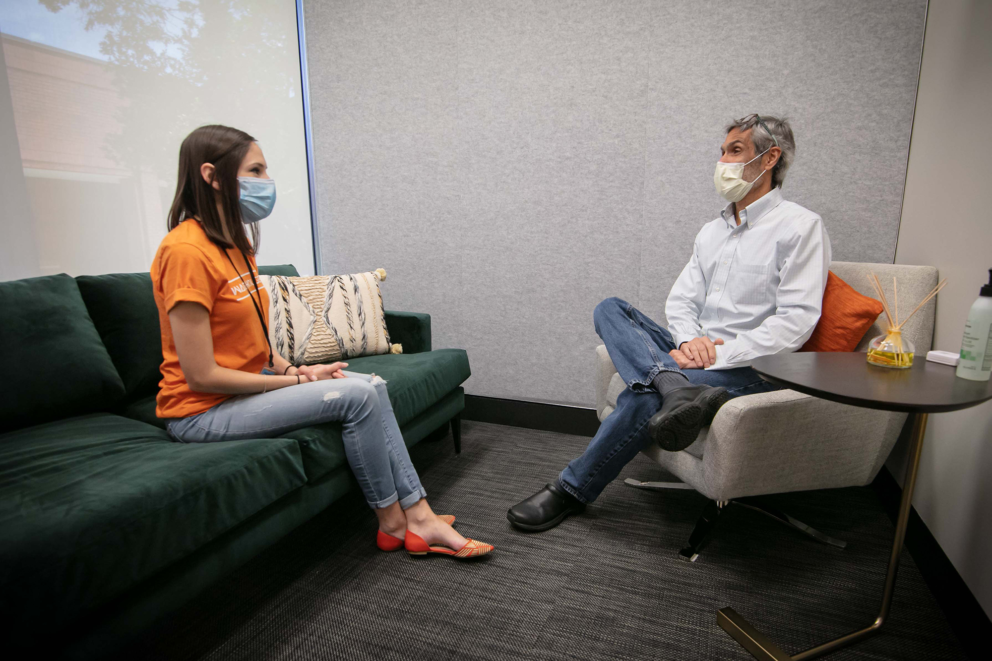 A man and a woman sit in a clinic setting wearing masks while speaking.