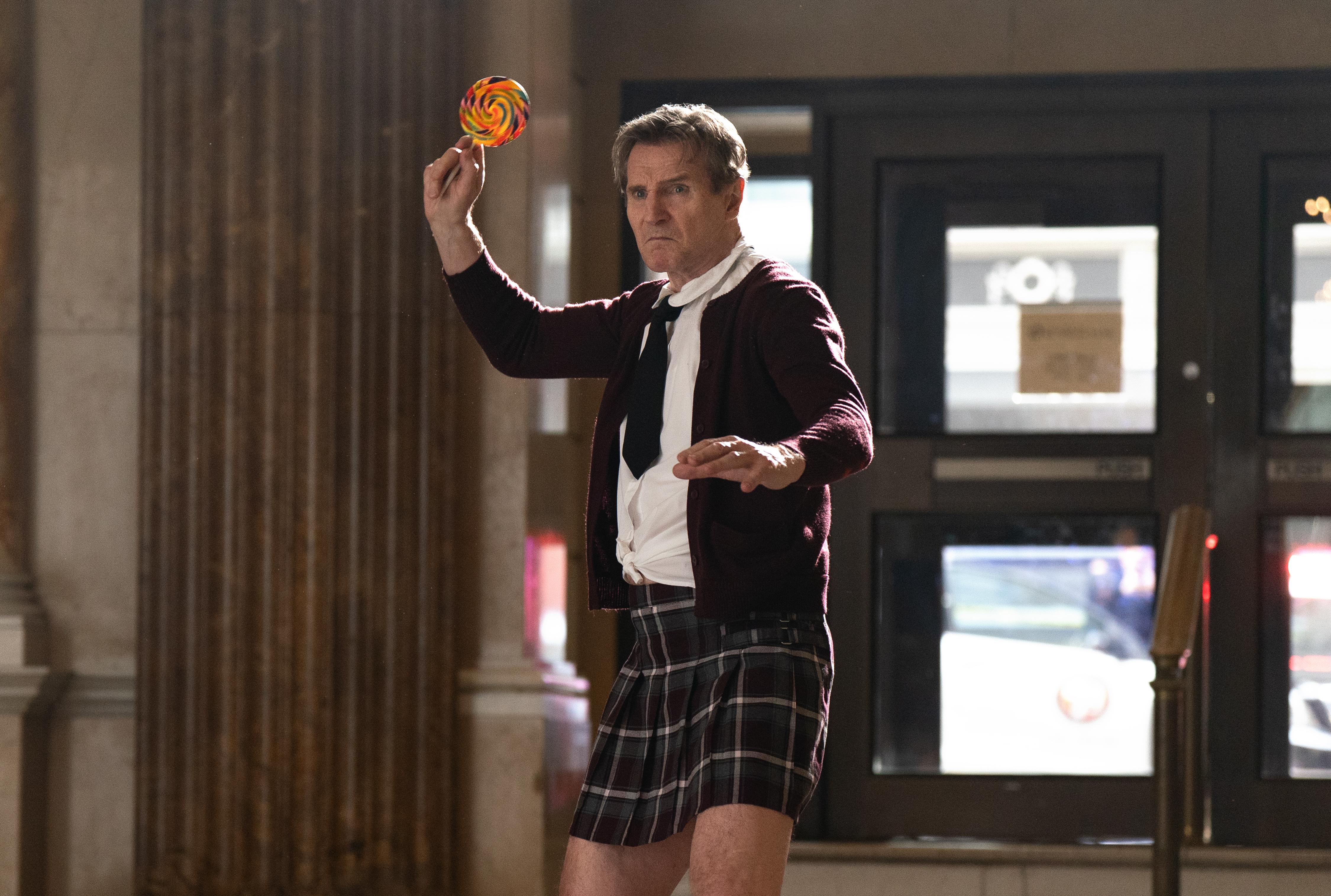 Liam Neeson wearing a girls school uniform and holding up candy