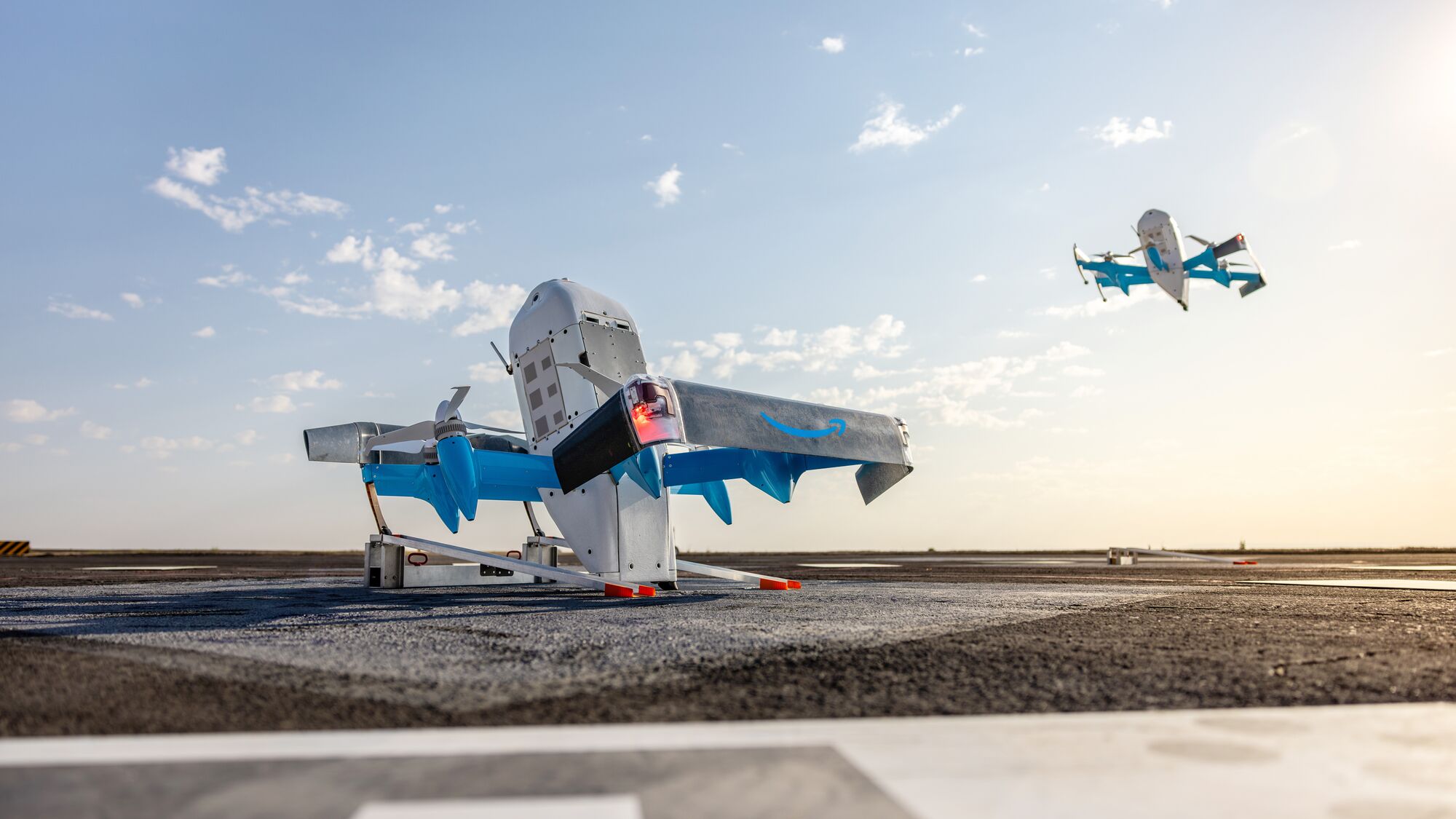 An exterior shot of two Prime Air MK30 delivery drones: One parked on the ground and one hovering in the sky to the right.