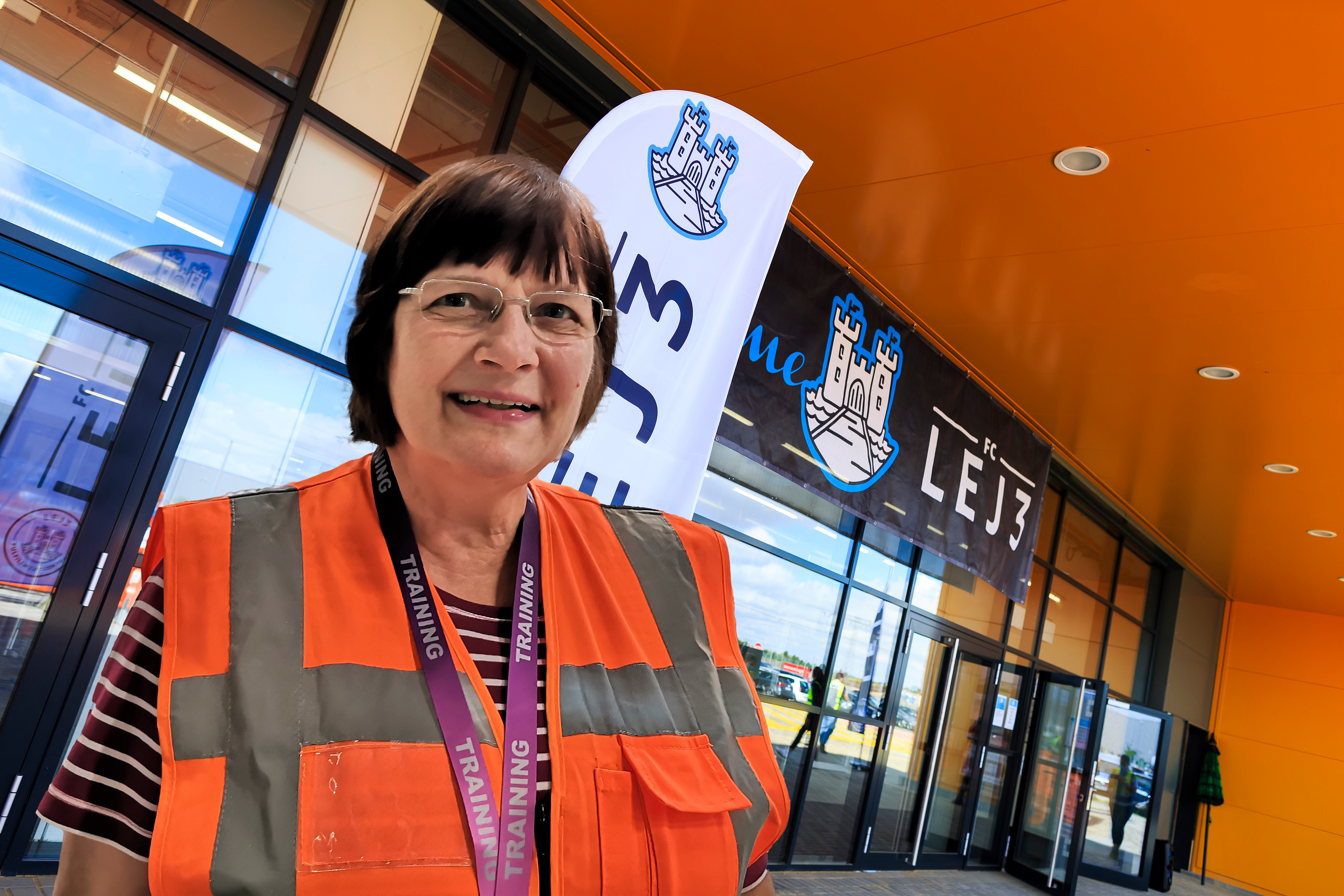 Eine Frau mit Brille und braunen Haaren, sie trägt eine orangene Sicherheitsweste. Im Hintergrund sieht man das Logistikzentrum.