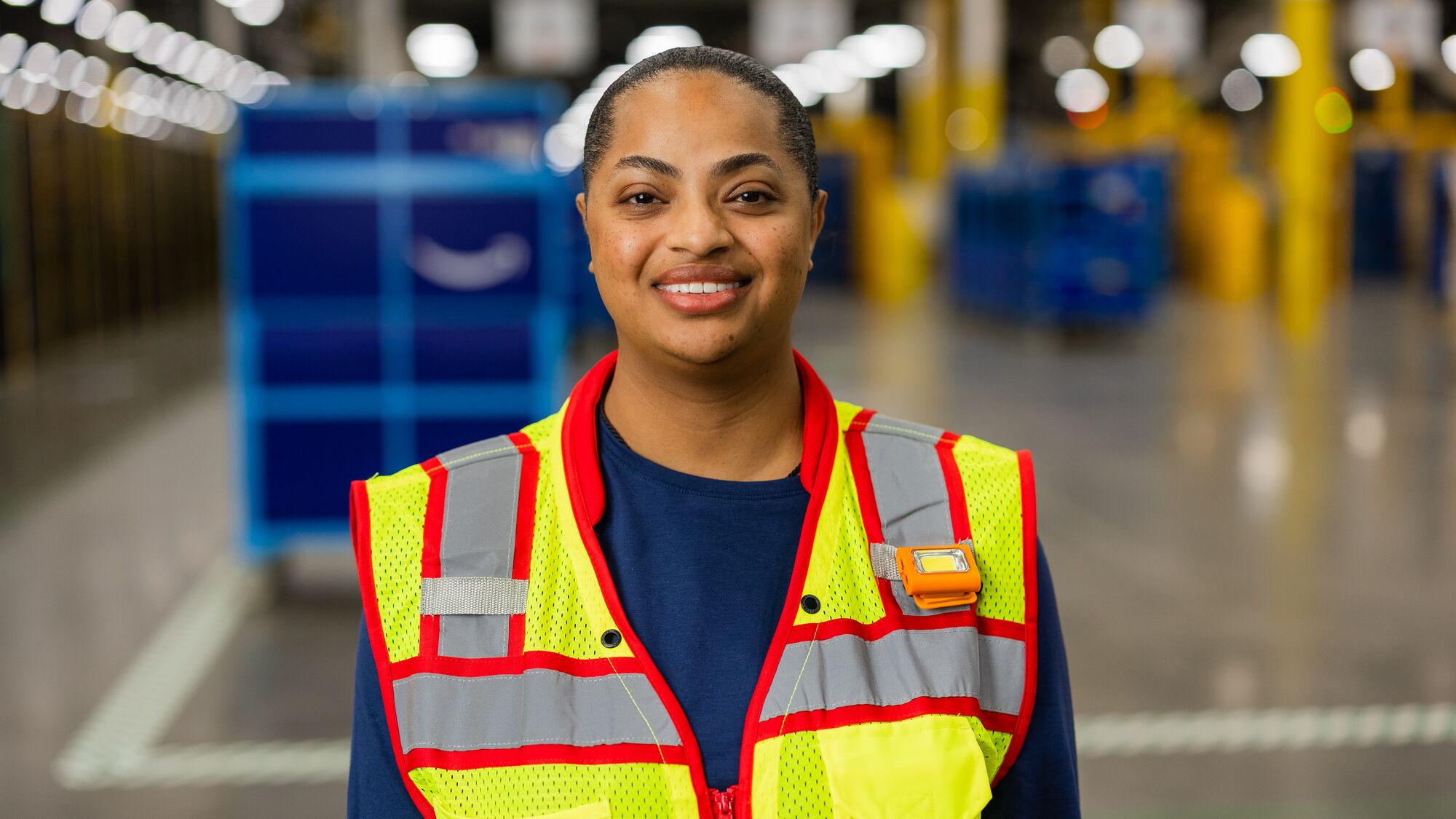 Amazon employee wears a safety vest and smiles while standing in a fulfillment center.