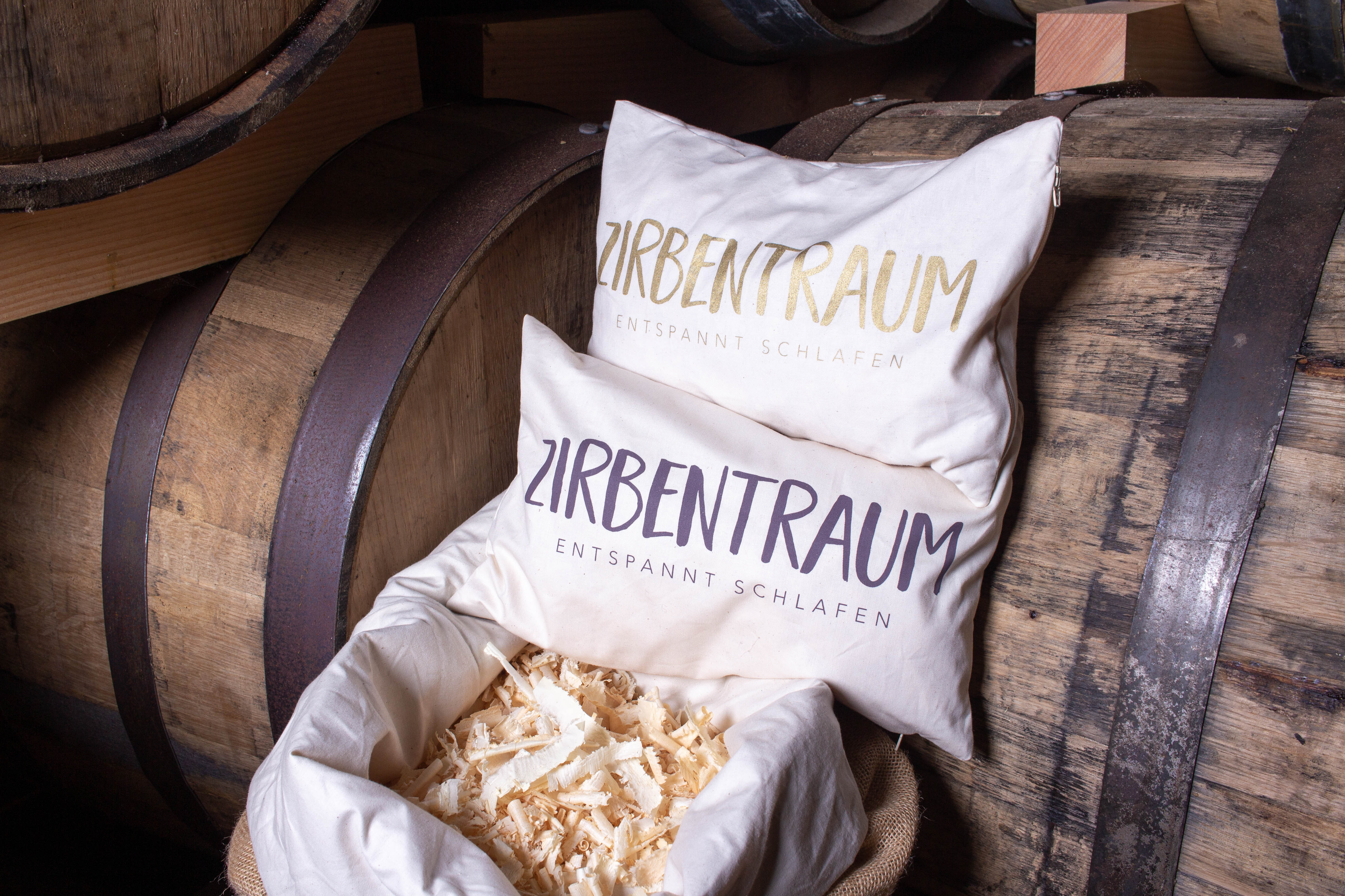 Zwei weiße Kissen mit der Aufschrift Zirbentraum liegen auf Holzfässern.