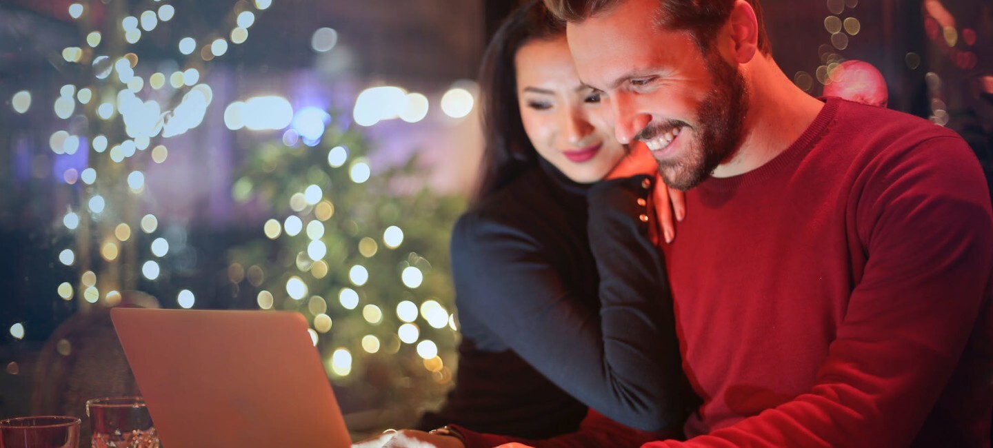 Ein Mann und eine Frau schauen lächelnd in einen hellgrauen Laptop. Im Hintergrund sind verschwommen Weihnachtsbäume und Lichter zu sehen.