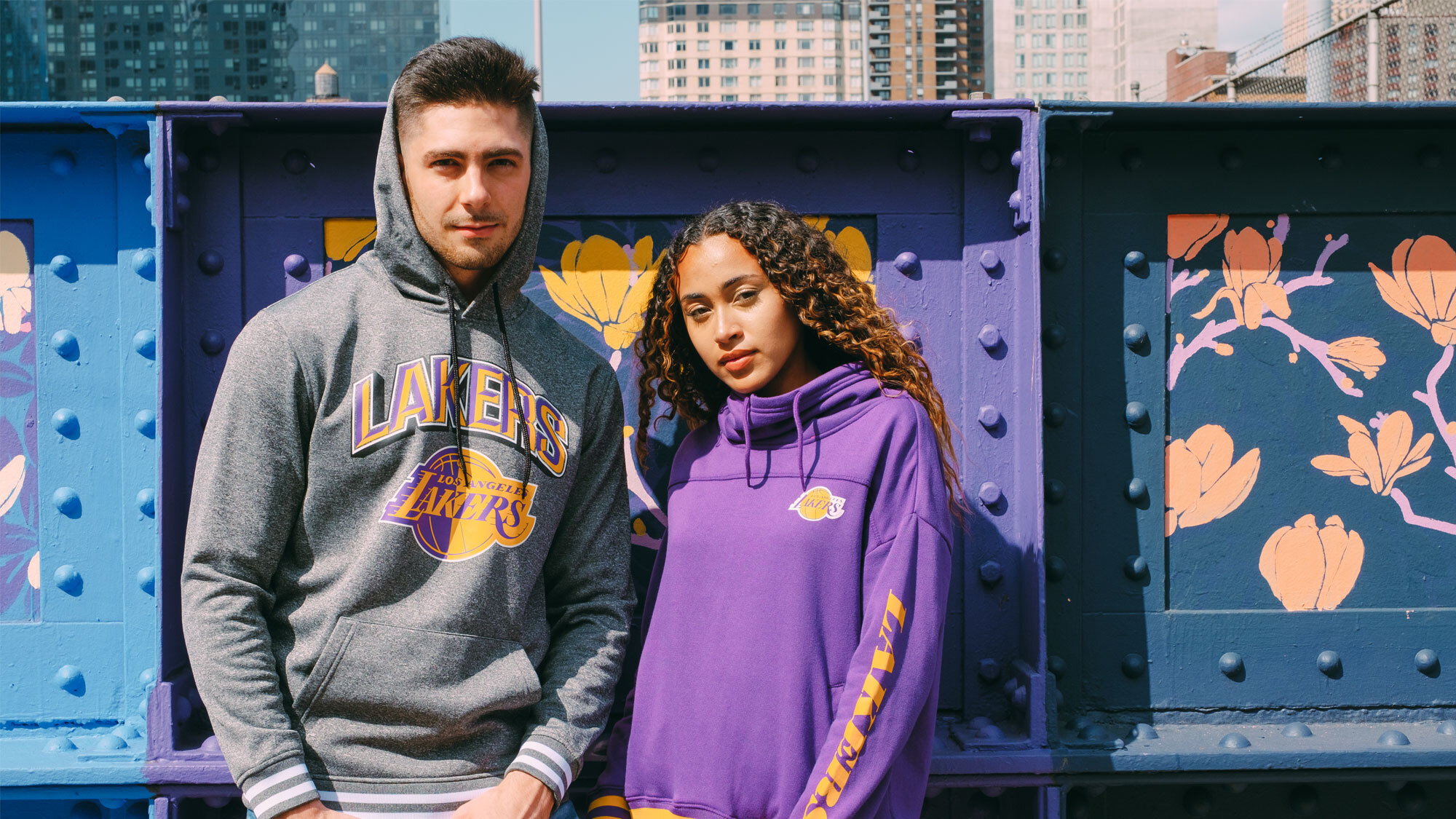 An image of a man and a woman standing outside in front of a metal wall with colorful, yellow, purple, and blue murals painted on it. The man is wearing a gray hoodie that says "Lakers" on it with a Lakers logo, and the woman is wearing a purple hoodie with the Lakers logo on the left chest and yellow text that says "Lakers" on the sleeve.