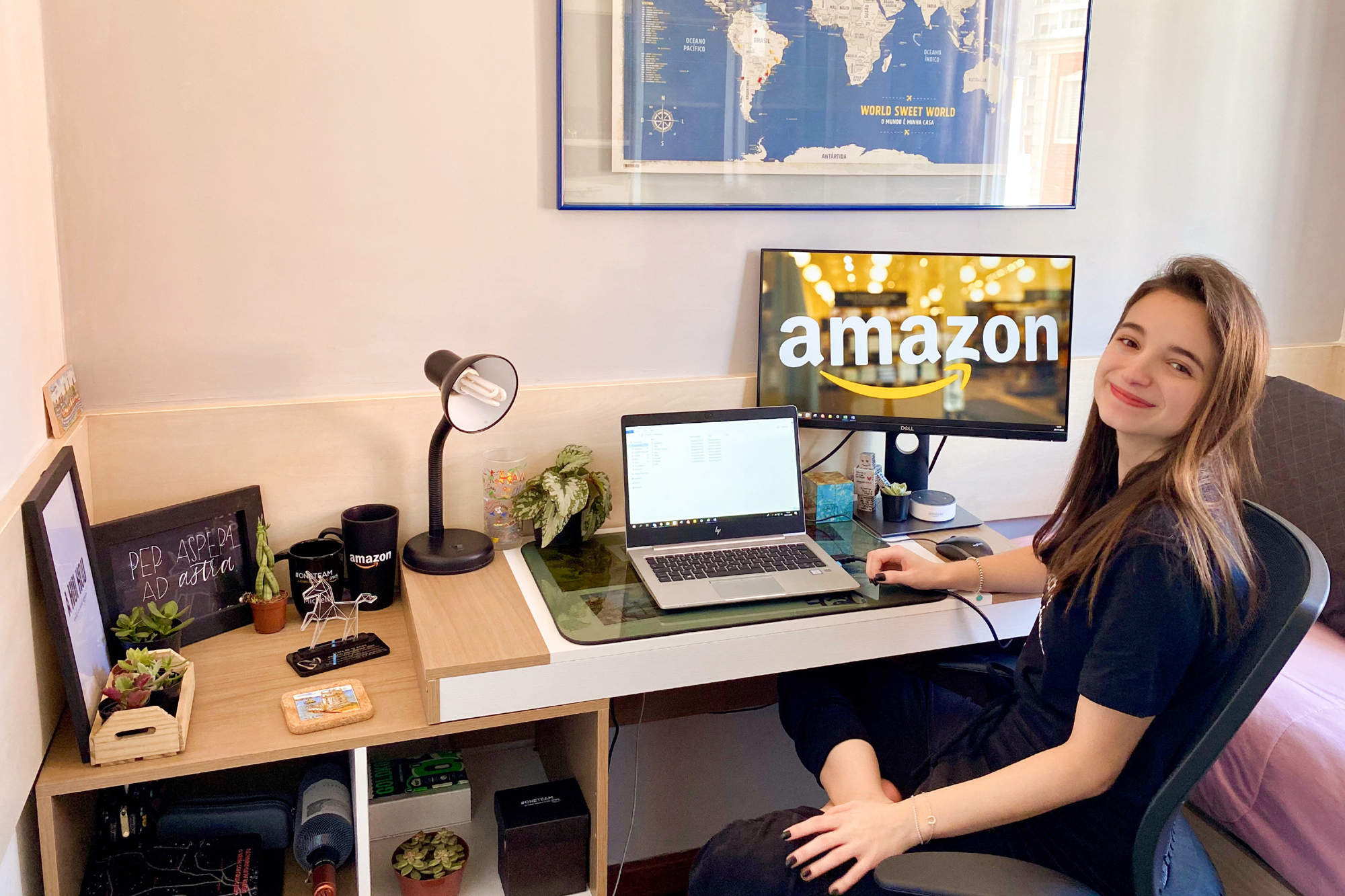 A woman sits at a desk with a laptop and a monitor showing the Amazon logo.