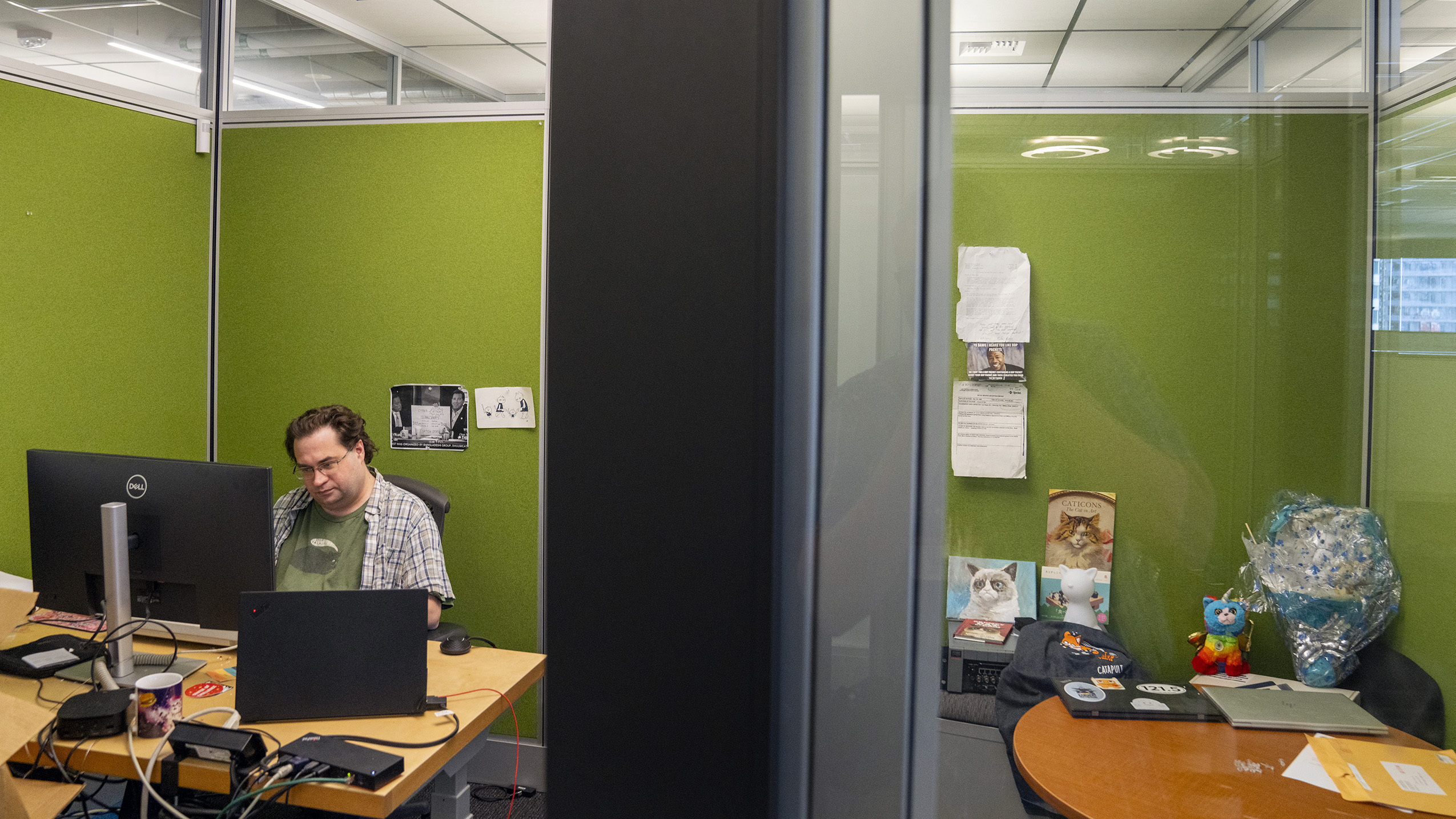 A photo of Tom Scholl, AWS VP and Distinguished Engineer, sitting at his desk, working on his computer. Hanging on the wall behind Tom are various printed graphics of cats.