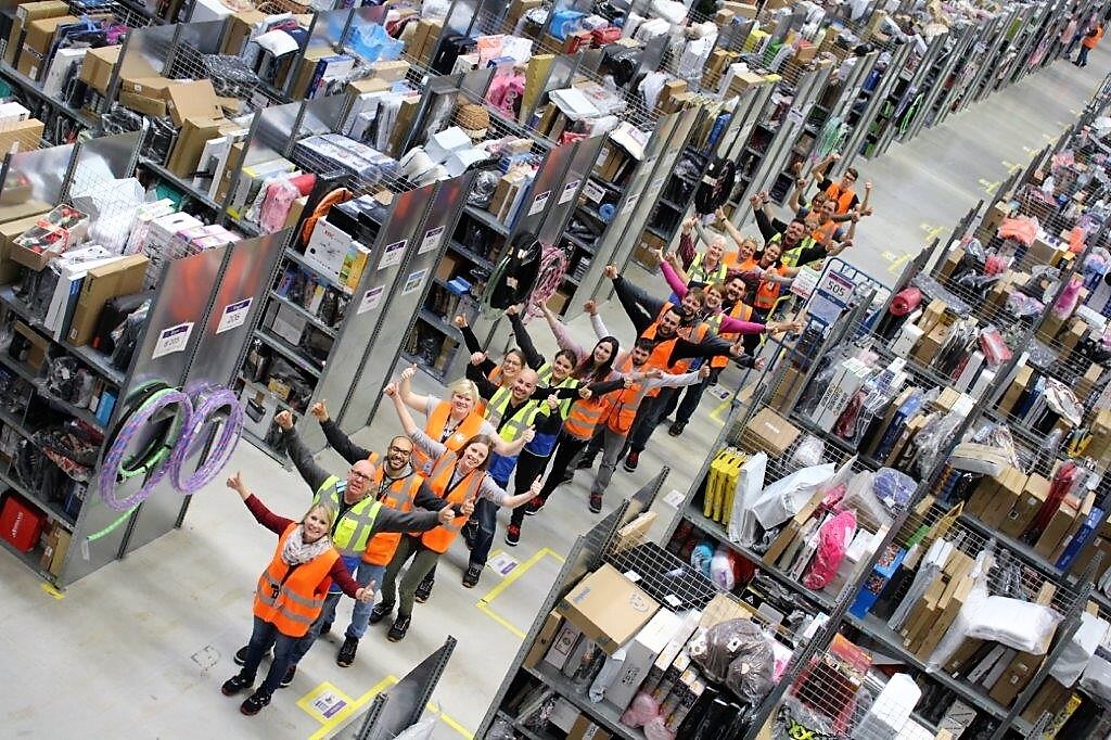 Eine Reihe von Logistikmitarbeitern stehen nacheinander im einem Regalgang, die Daumen nach oben
