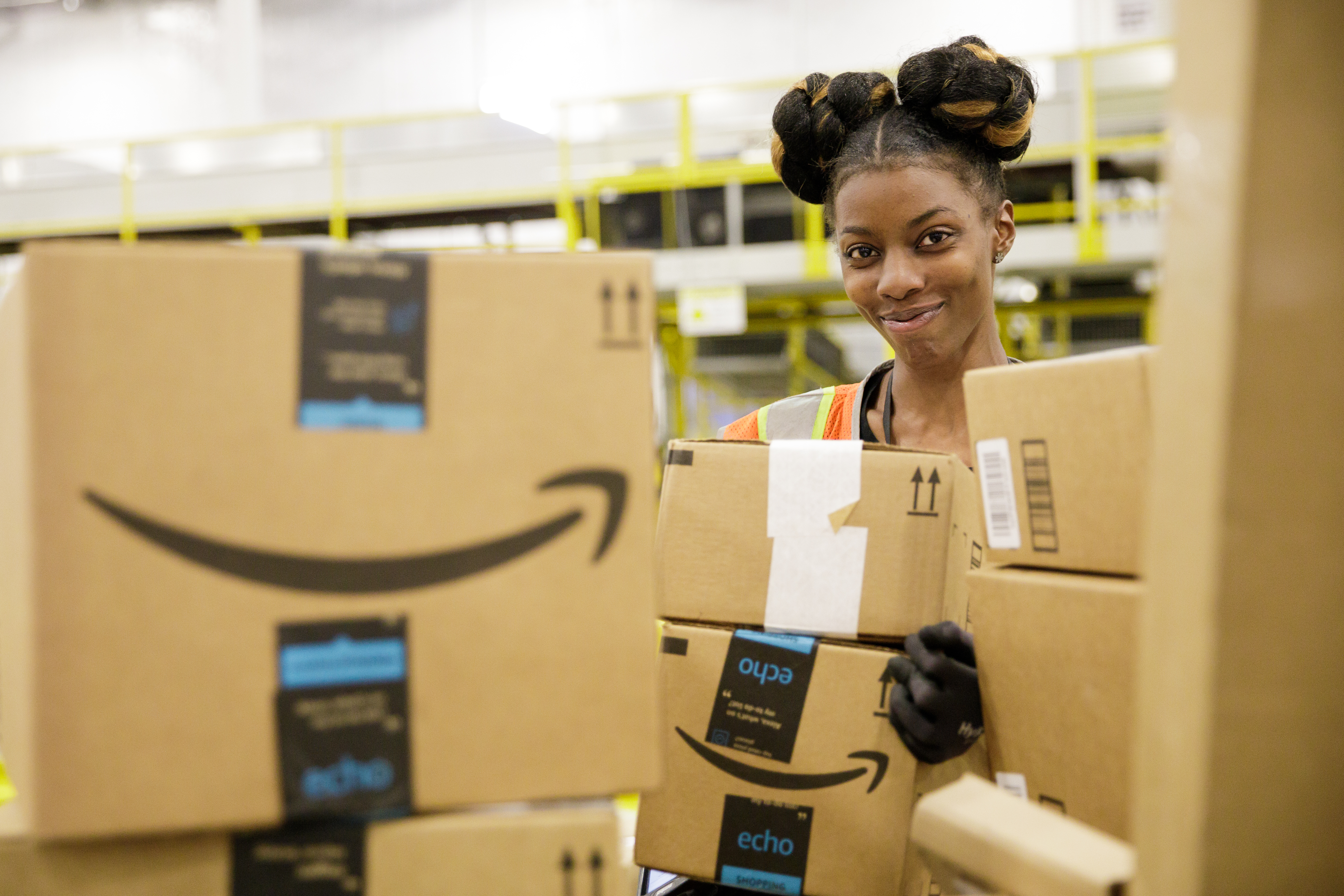 A smiling Amazon sortation associate in our Baltimore, Maryland sortation center looks at the camera from behind a pile of Amazon delivery boxes.