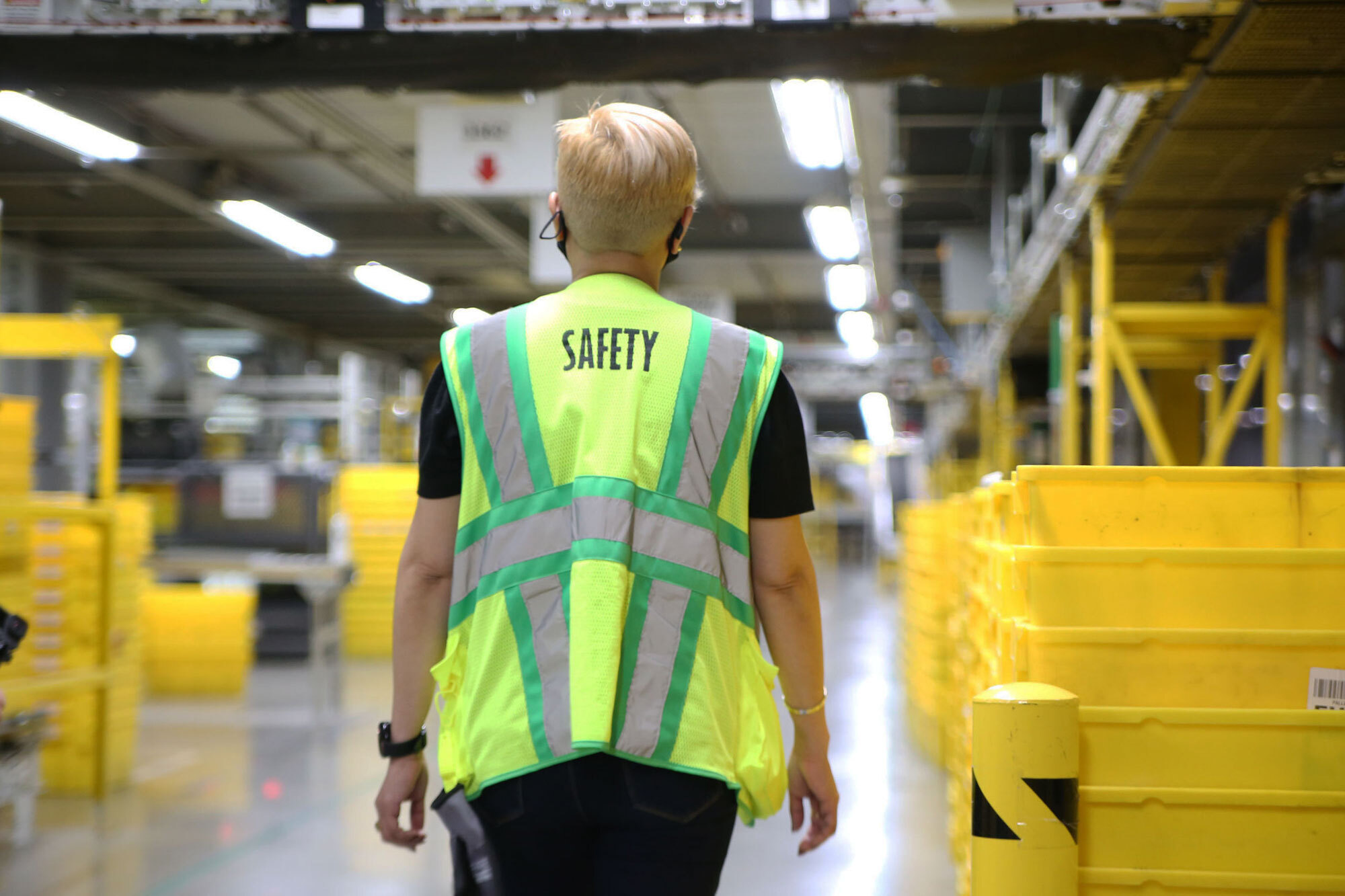 A woman with a safety vest walks through an Amazon fulfillment center with her back to the camera.