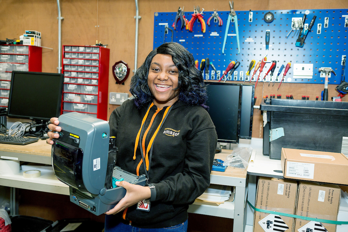 Apprentice, Shannia Daley, pictured in the workshop in an Amazon fulfilment centre holding an open printer machine and smiling.
