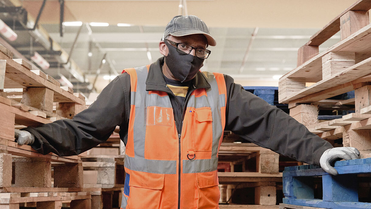 Amazon associate Melvyn wears a mask, high visibility vest, and safety gloves while standing amongst stacks of pallets on an Amazon fulfillment center shipping dock