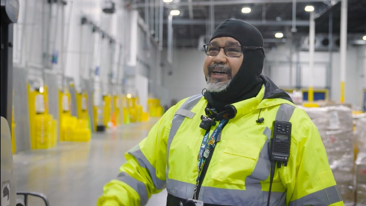 An Amazon employee smiles and wears a bright yellow safety jacket as he works in a fulfillment center.