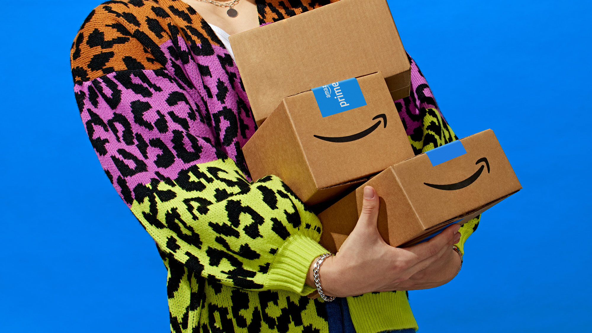 Prime Day 2024: Eine Person in bunter Weste, deren Kopf man nicht sieht, trägt  3 aufeinander gestapelte Amazon Päckchen zum Prime Day 2024, das Bild hat einen blauen Hintergrund