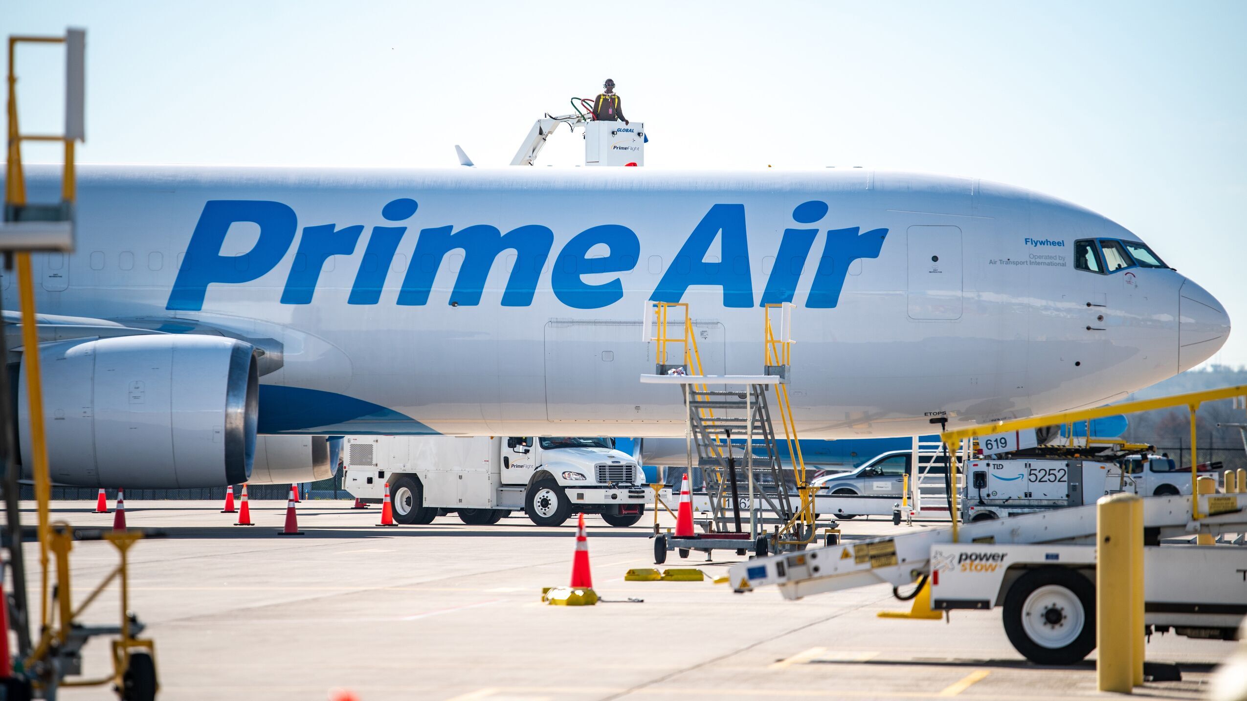 An image of an Amazon Air plane parked outside of an Amazon Air hub. There are various vehicles parked around the plane with employees working on different areas. 