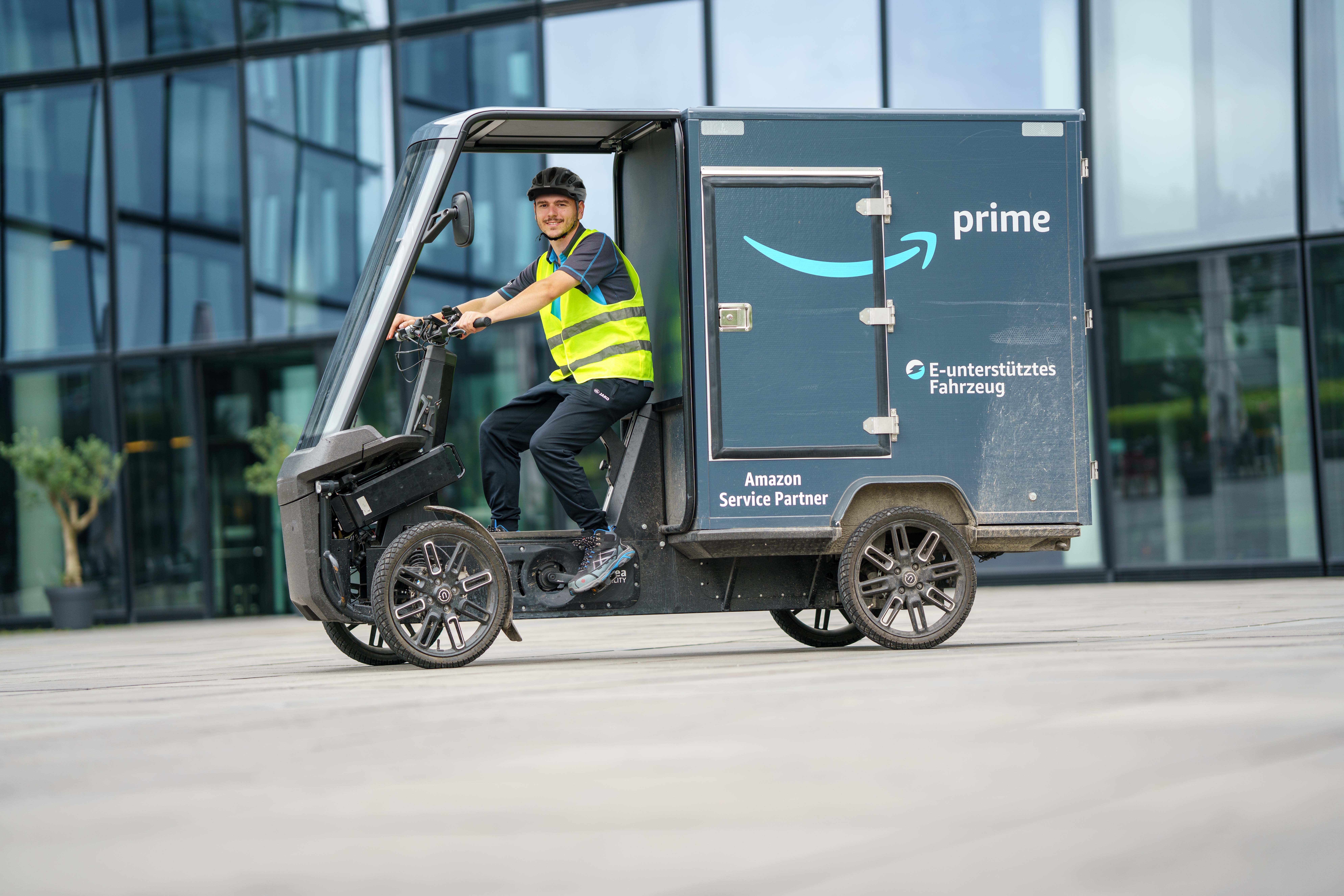 Ein junger Amazon-Fahrer mit dem E-Cargo Bike: Er trägt Berufskleidung im Amazon-look und ist mit einem blau-grauen E-Lastenrad unterwegs.