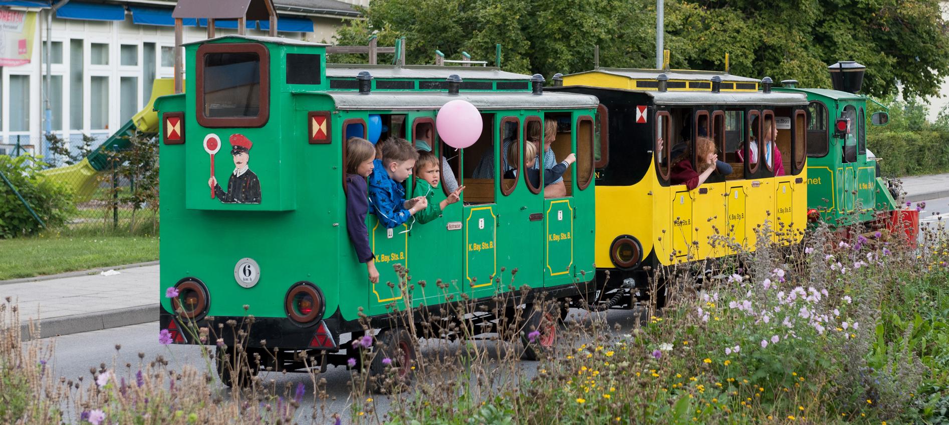 Kinder fahren in einer kleinen Lokomotive auf der Straße. Im Hintergrund ist ein Spielplatz, das Krankenhaus und eine Blumenwiese. Die Kinder schauen aus den Fenstern der Wagons und haben Spaß.