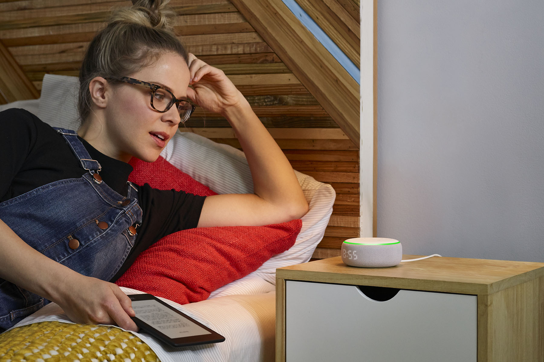 Woman wearing glasses lays on a bed as she looks at and speaks to an Amazon Echo device on a nightstand. 