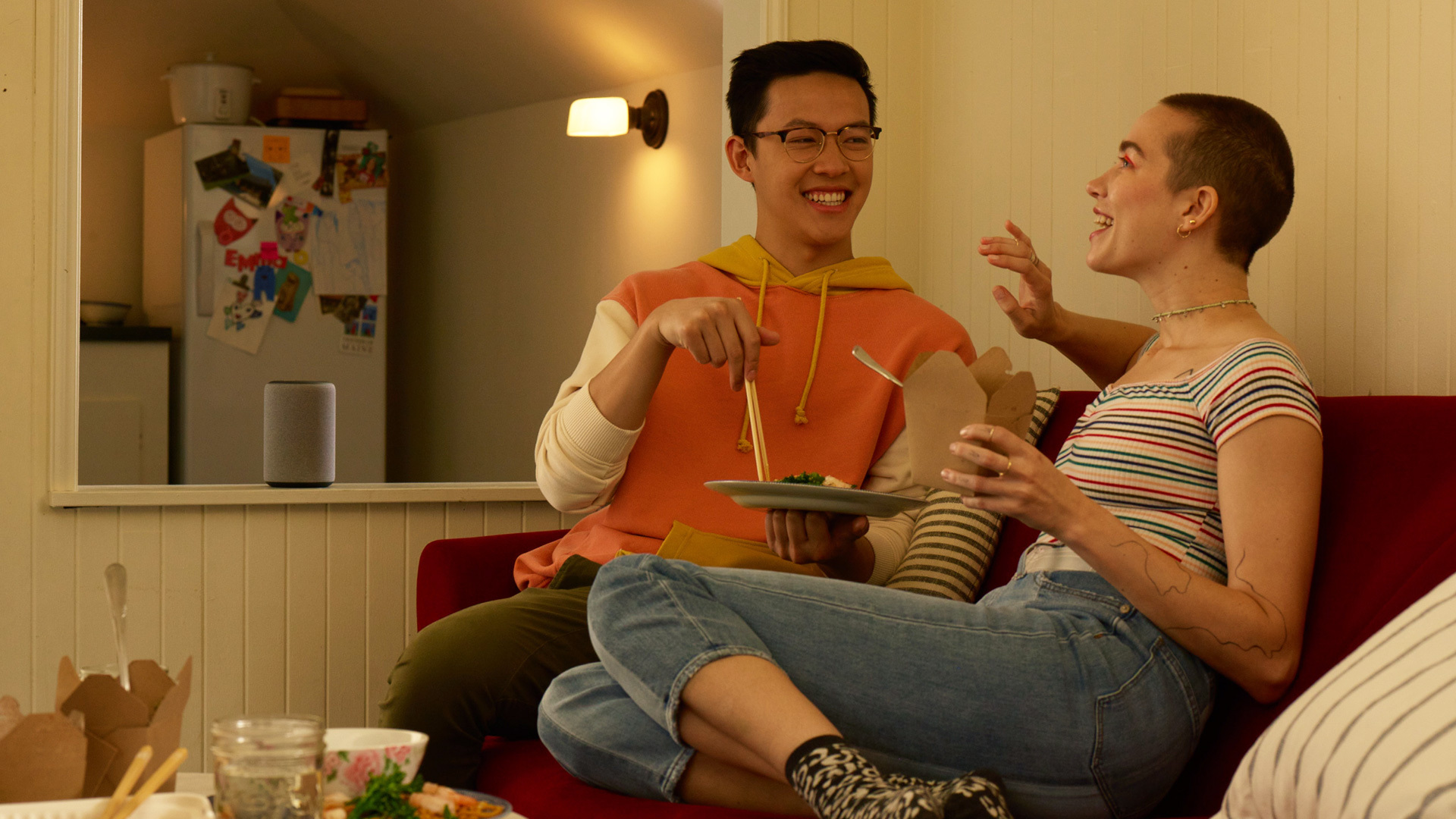 An image of two people sitting in a sofa having a conversation with plates of food in their hands. Behind them is a kitchen and an Amazon Echo device on the counter. 