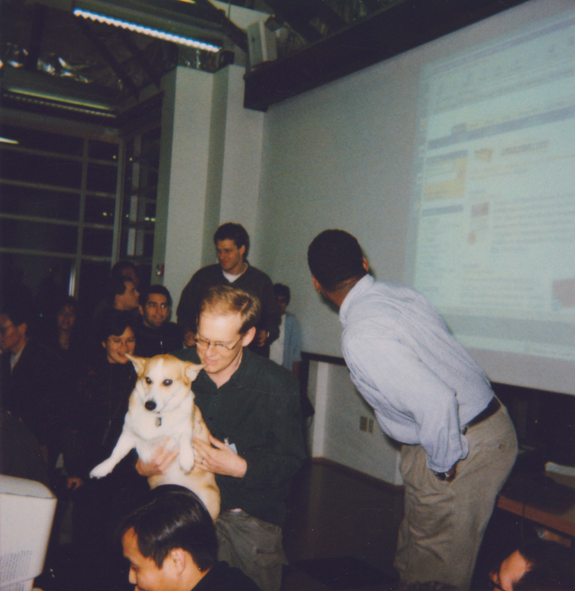 A Welsh Corgi named Rufus, the first dog of Amazon, is being held in a man's lap. His paw is poised to launch a new site. Round him are other early Amazonians. 