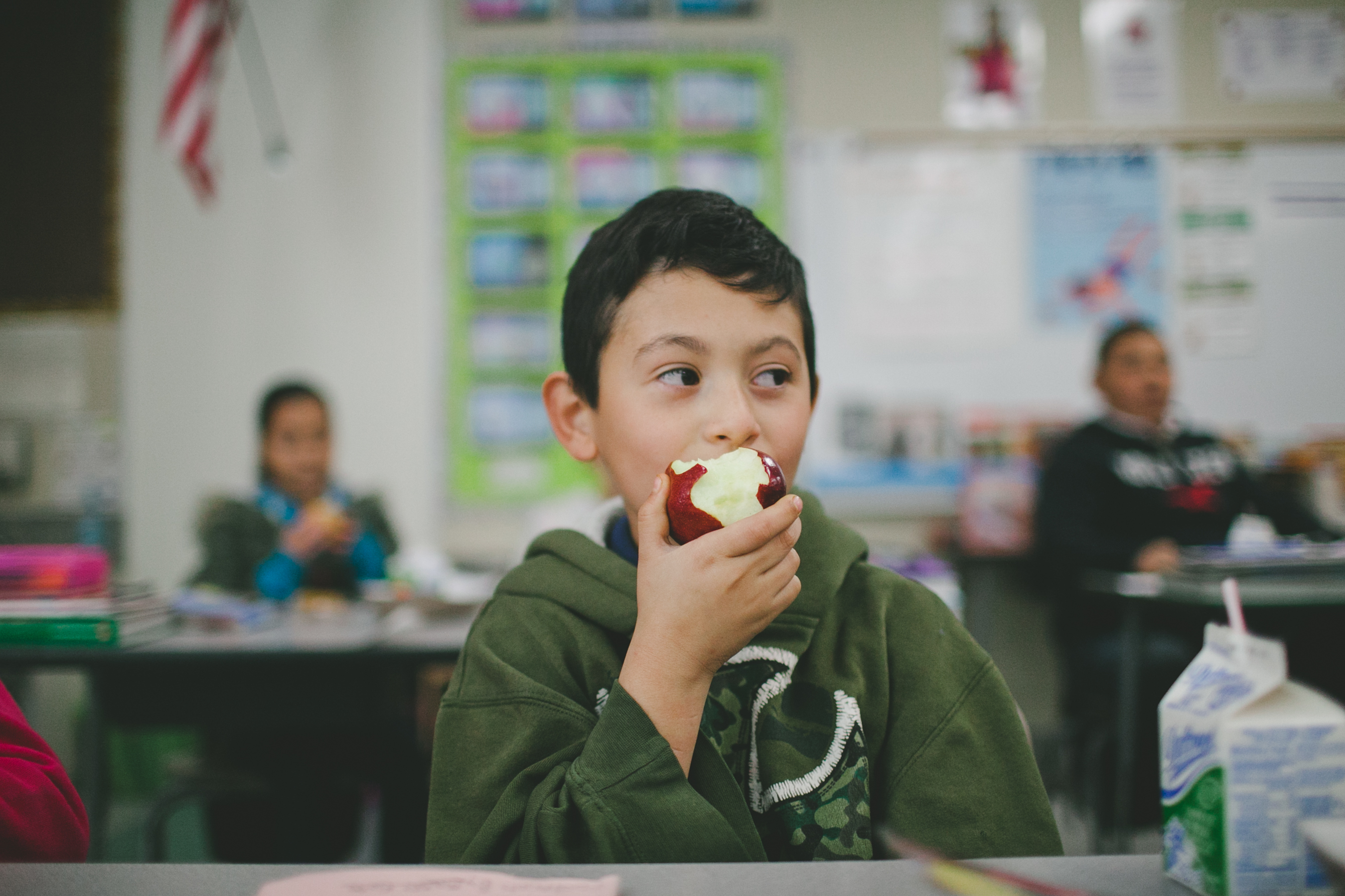 A boy in a classroom is eating an apple. Behind him, other students are seated at desks.