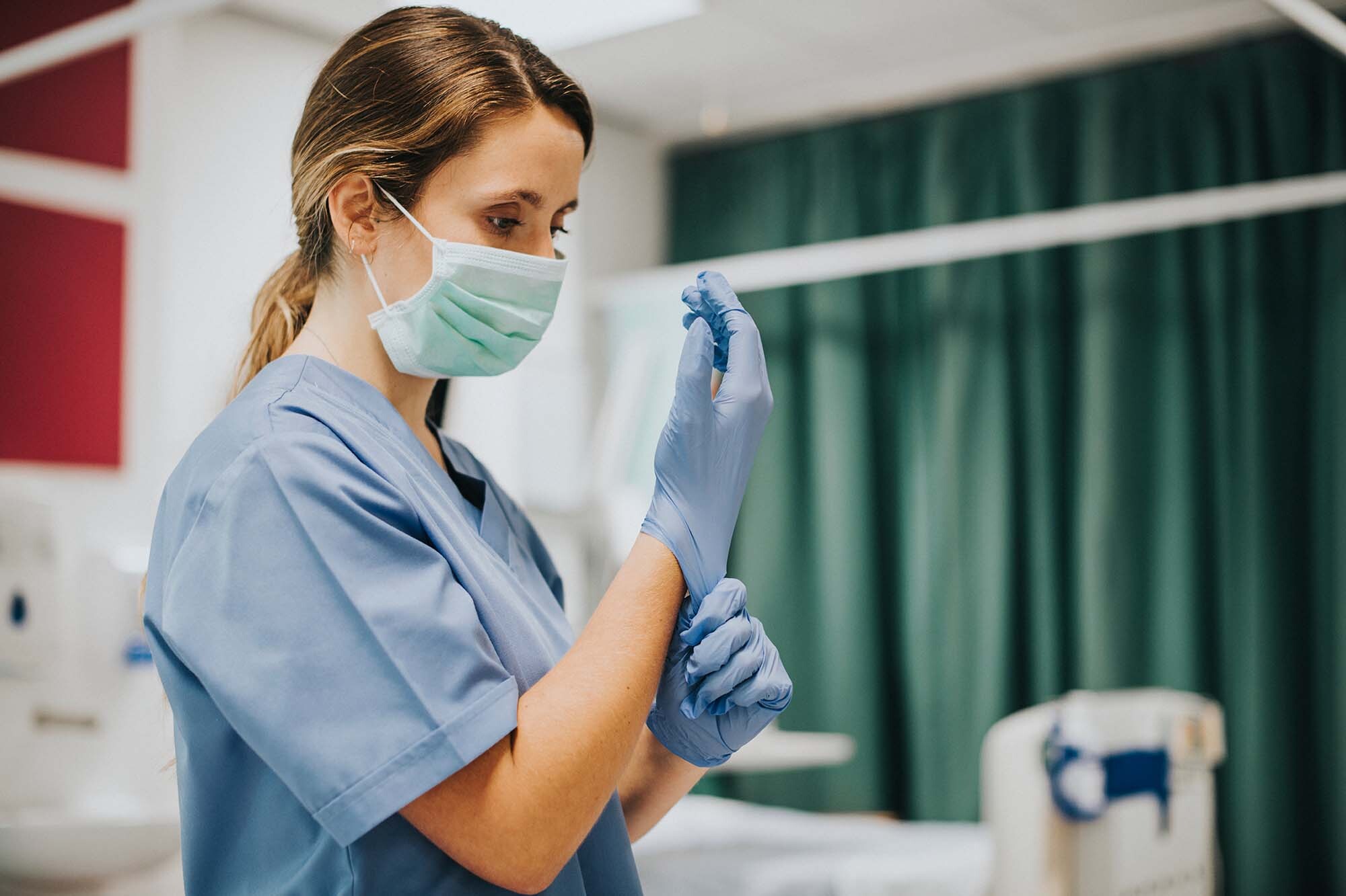 A woman wearing a face mask and scrubs pulls on gloves in a medical setting.