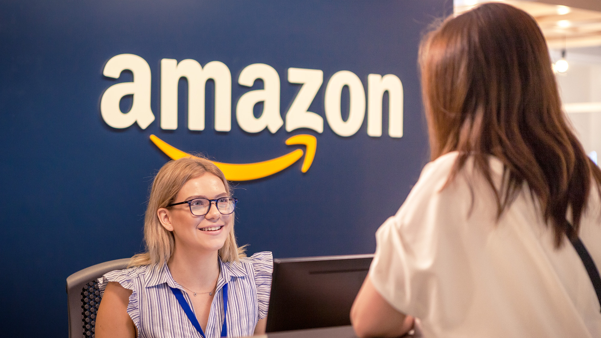 An image of a woman in the front desk of an Amazon office greeting another woman. There is an amazon logo on the wall behind her chair. 