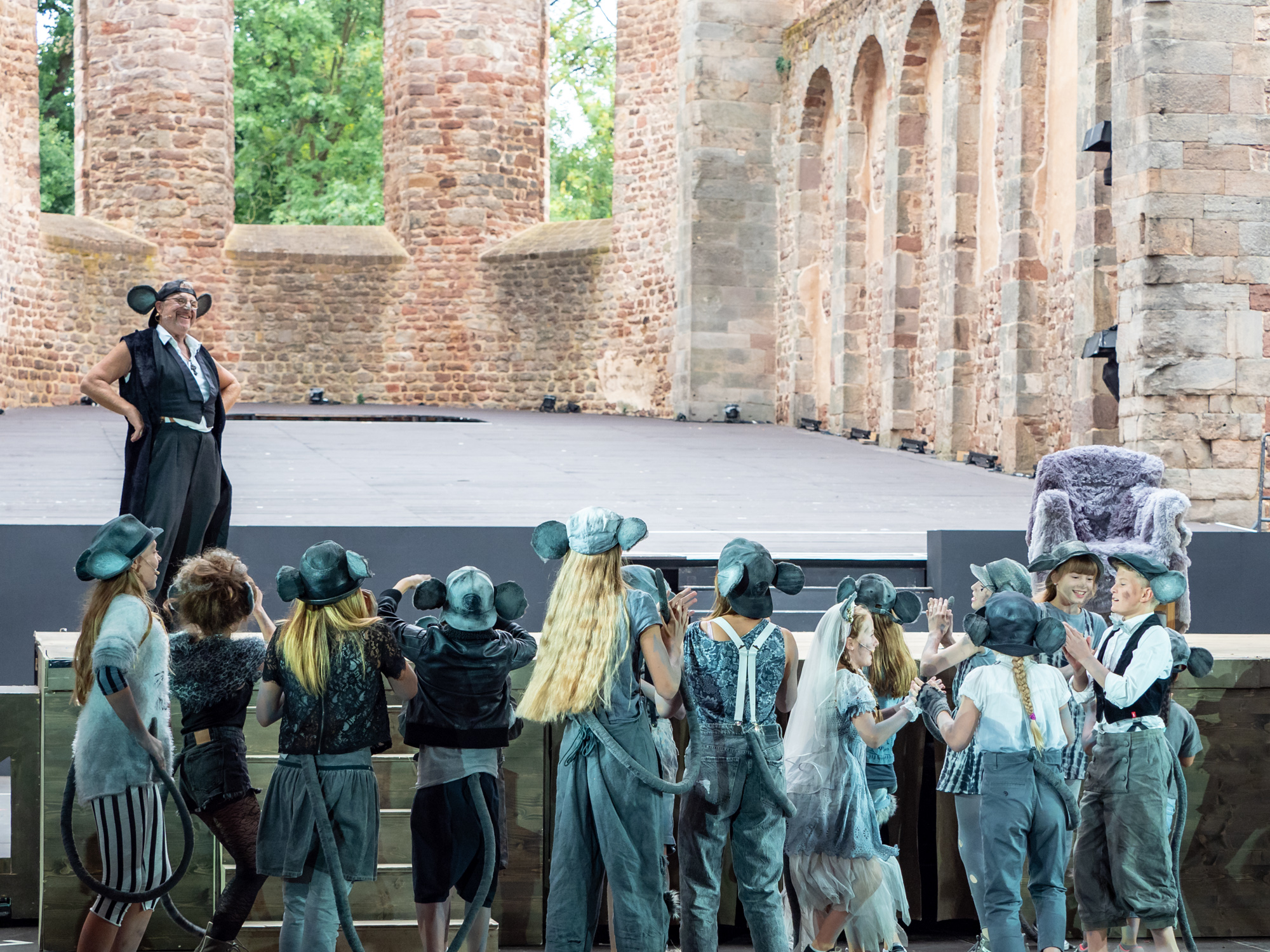In der historischen Stiftsruine Bad Hersfeld ist eine große Bühne aufgebaut, im Hintergrund sieht man Mauerwerk in Backsteinen und glaslose Fenster der ehemaligen Stiftsruine. Auf der Bühne ein Schauspieler im Mäusekostüm. Vor ihm stehen ca. ein Dutzend Kinder, ebenfalls in Mäusekostümen. 