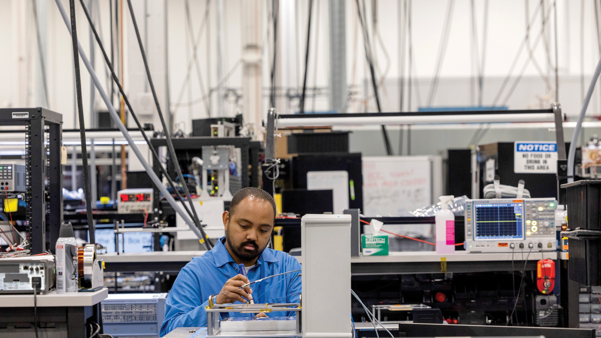 Man in blue lab coat sits at a workbench inside of an Amazon Project Kuiper factory.