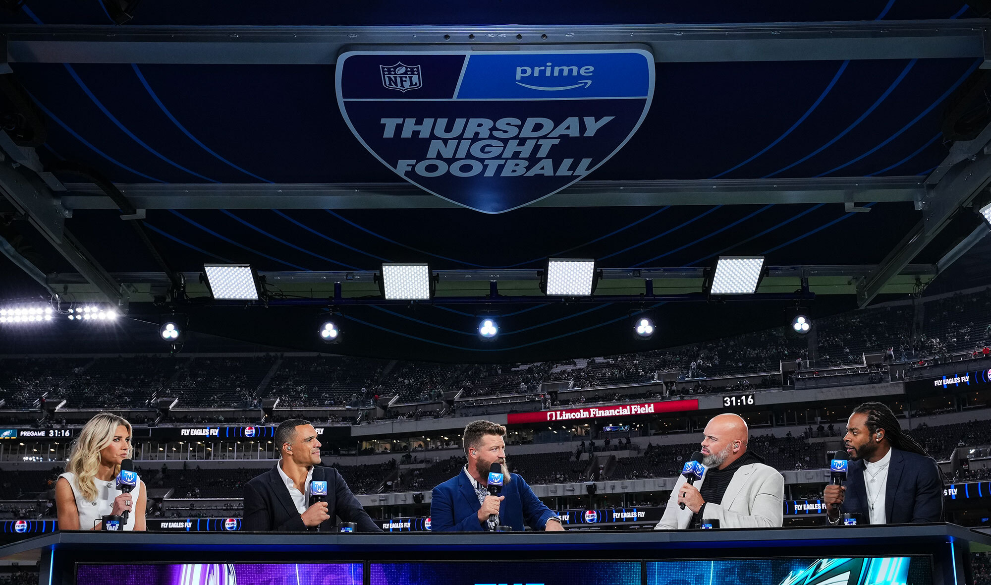 An image of Thursday Night Football announcers, reporters, and hosts in front of an NFL football field. They are standing around a desk and smiling while holding microphones.