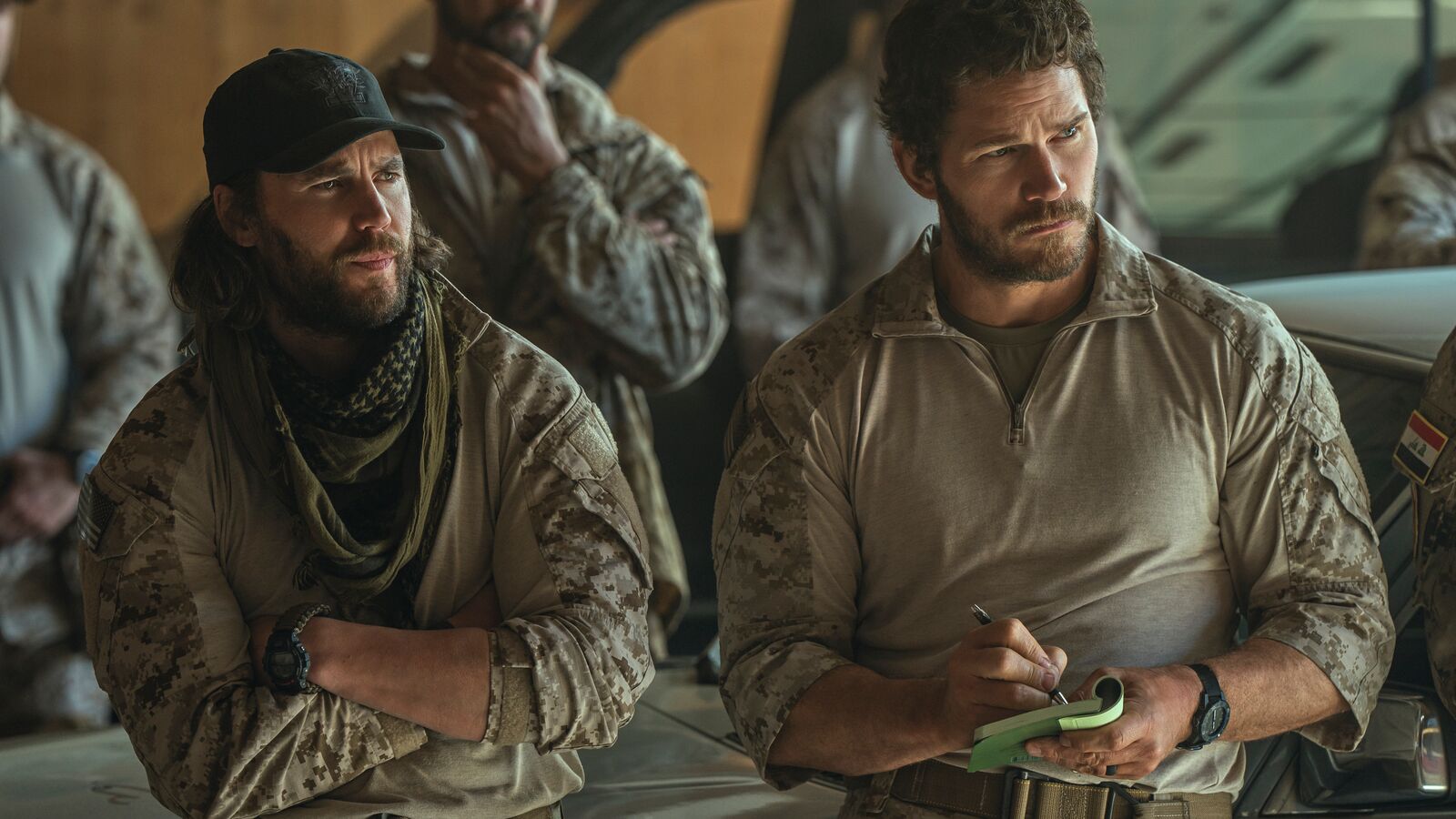 Two military personnel in desert camouflage uniforms looking serious.