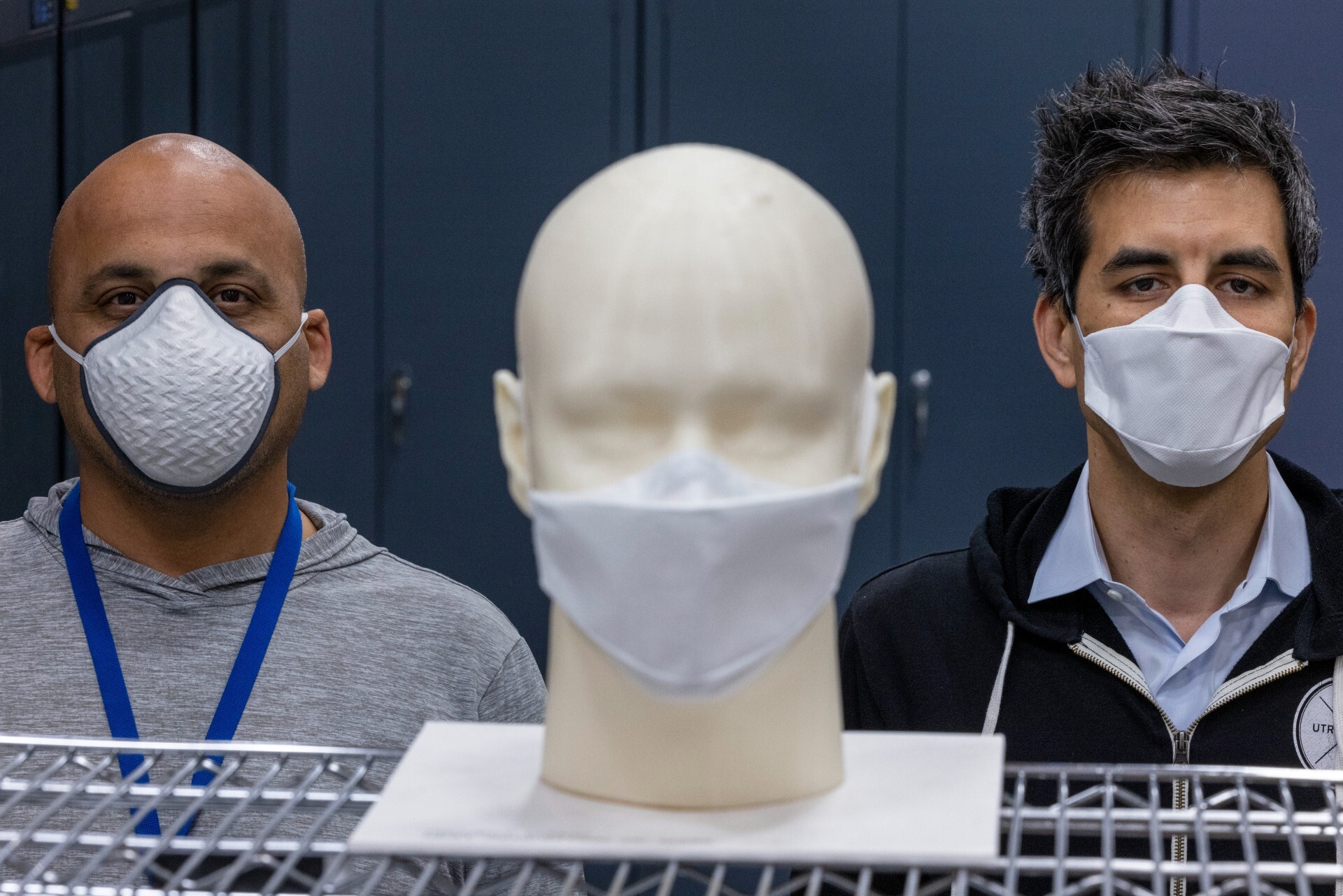 Two men wearing face masks stand behind a mask prototype.