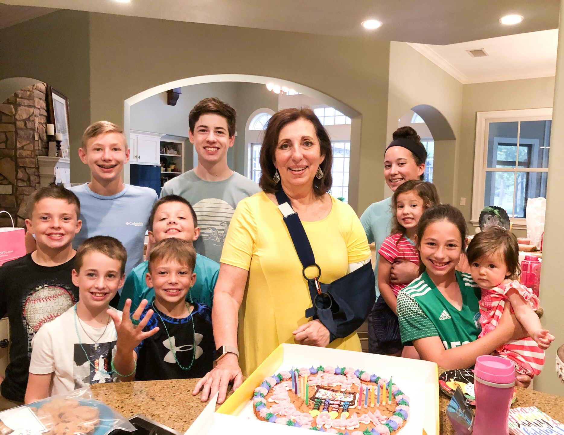 Rebecca in a sling supporting her left arm is surrounded by her ten grandchildren with a birthday cake in front of them.