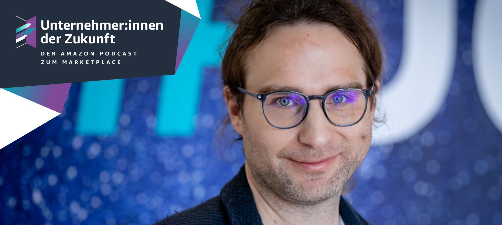 Ein Mann mit braunen langen Haaren und einer Brille lächelt in die Kamera.
