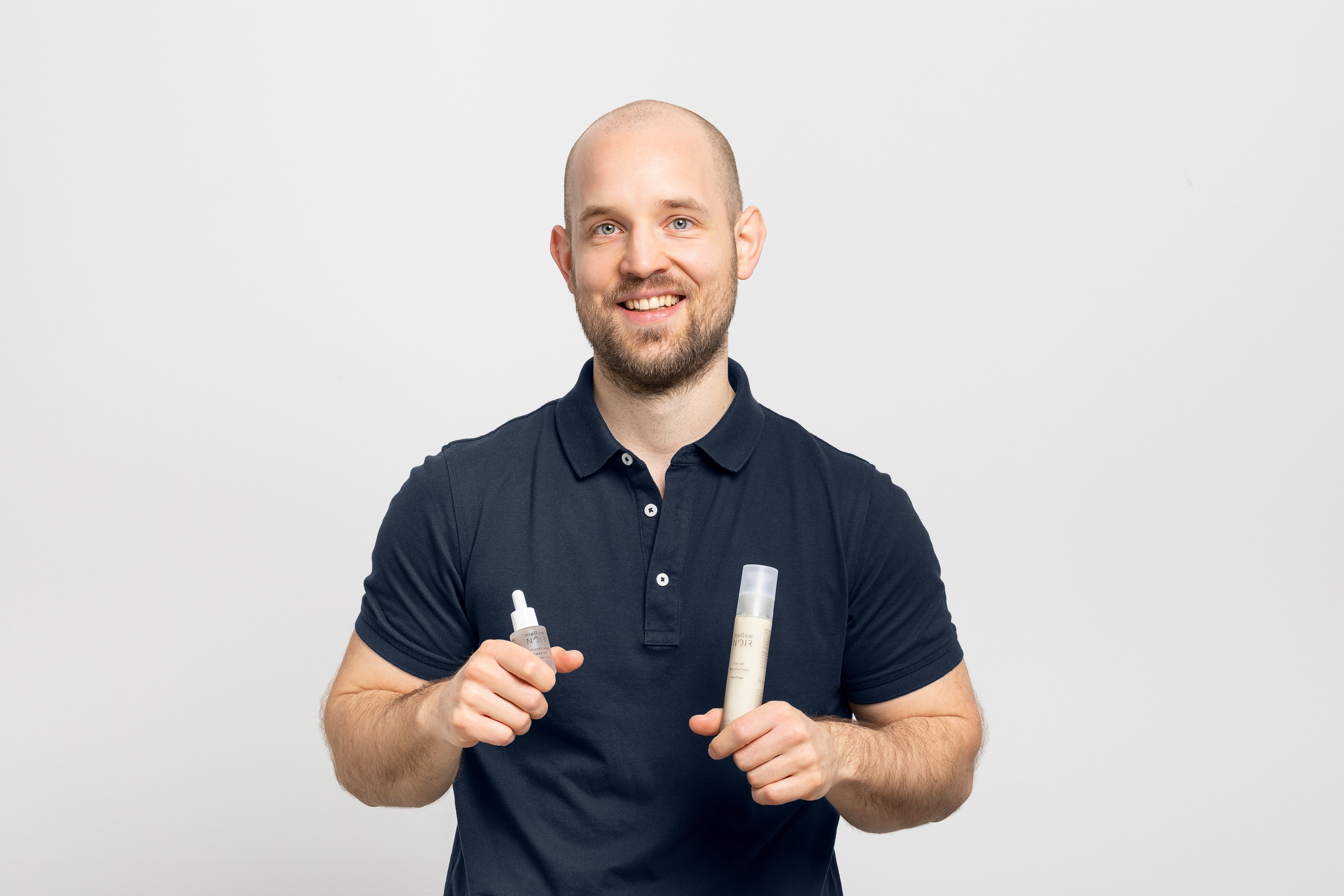 Ein Mann mit Glatze und einem schwarzen Poloshirt lacht in die Kamera. In seinen Händen hält er Produkte von Mellow Noir.