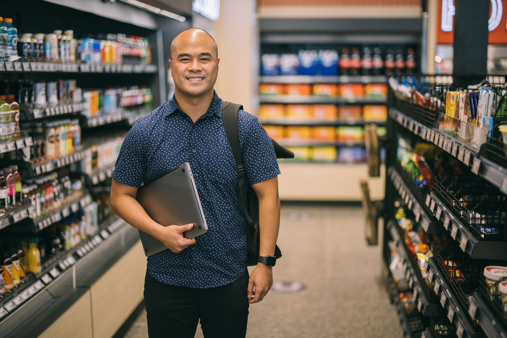 A man holding a laptop computer in one arm smiles at the camera from within an Amazon Fresh grocery store.