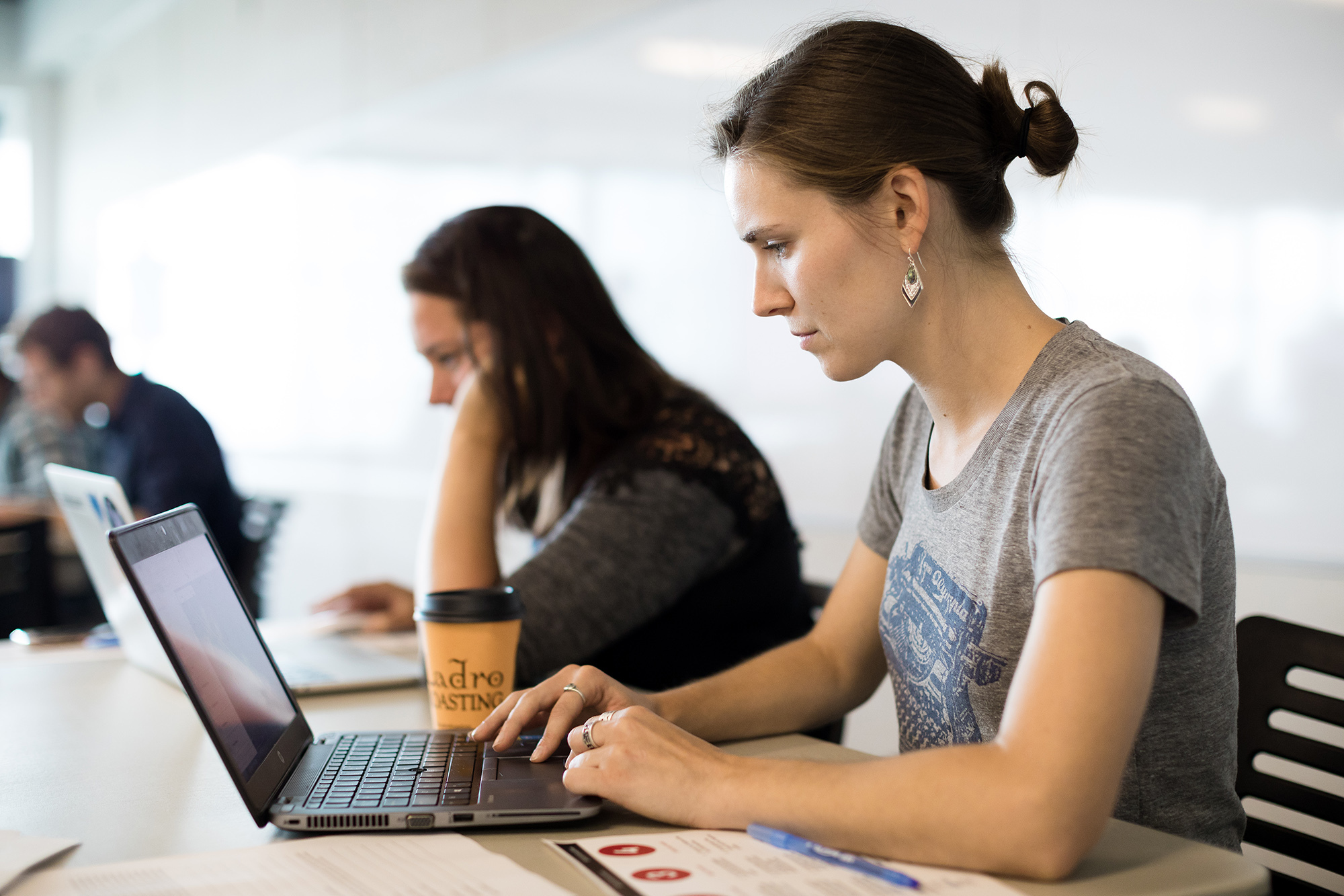 A woman in a t-shirt works on a laptop computer in a workplace setting. Next to her, another woman works on a laptop, and behind them, men sit at a table. 