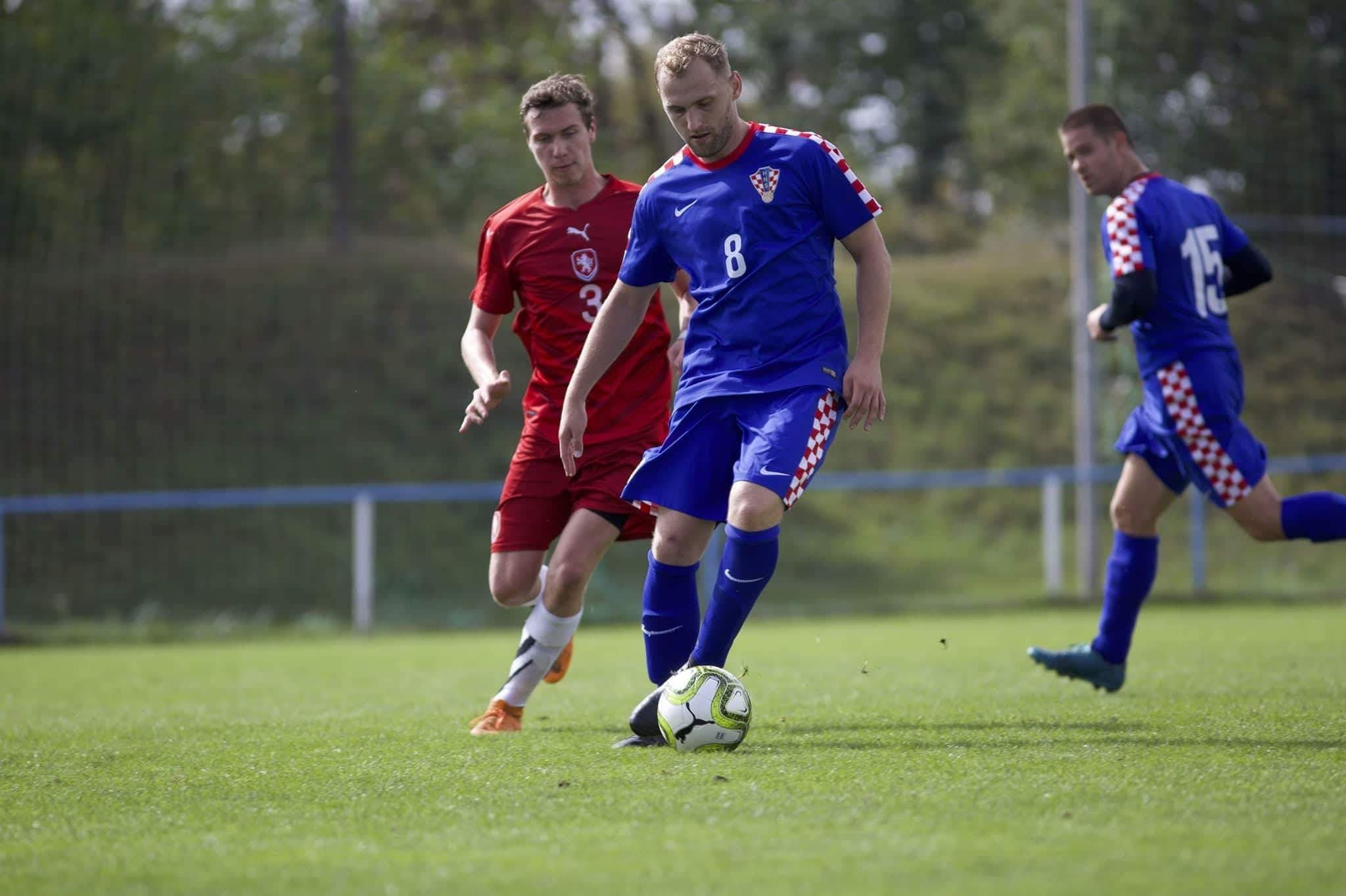 Zwei Fußballspieler auf einem Fußballfeld: Einer trägt ein rotes Trikot, der andere ist in blau. Beide springen hoch.