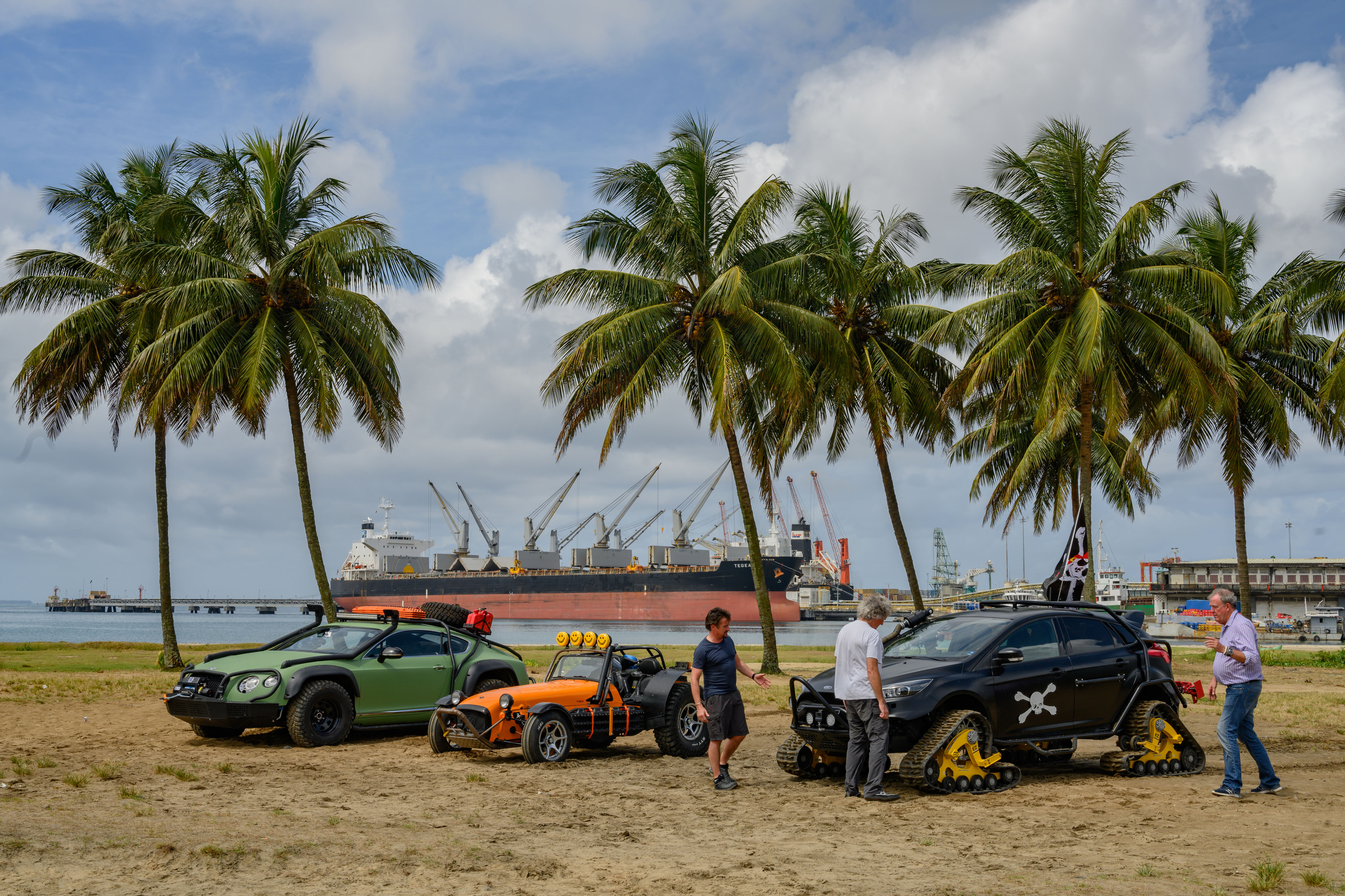 Three men stand next to three cars on a beach with palm trees in the background